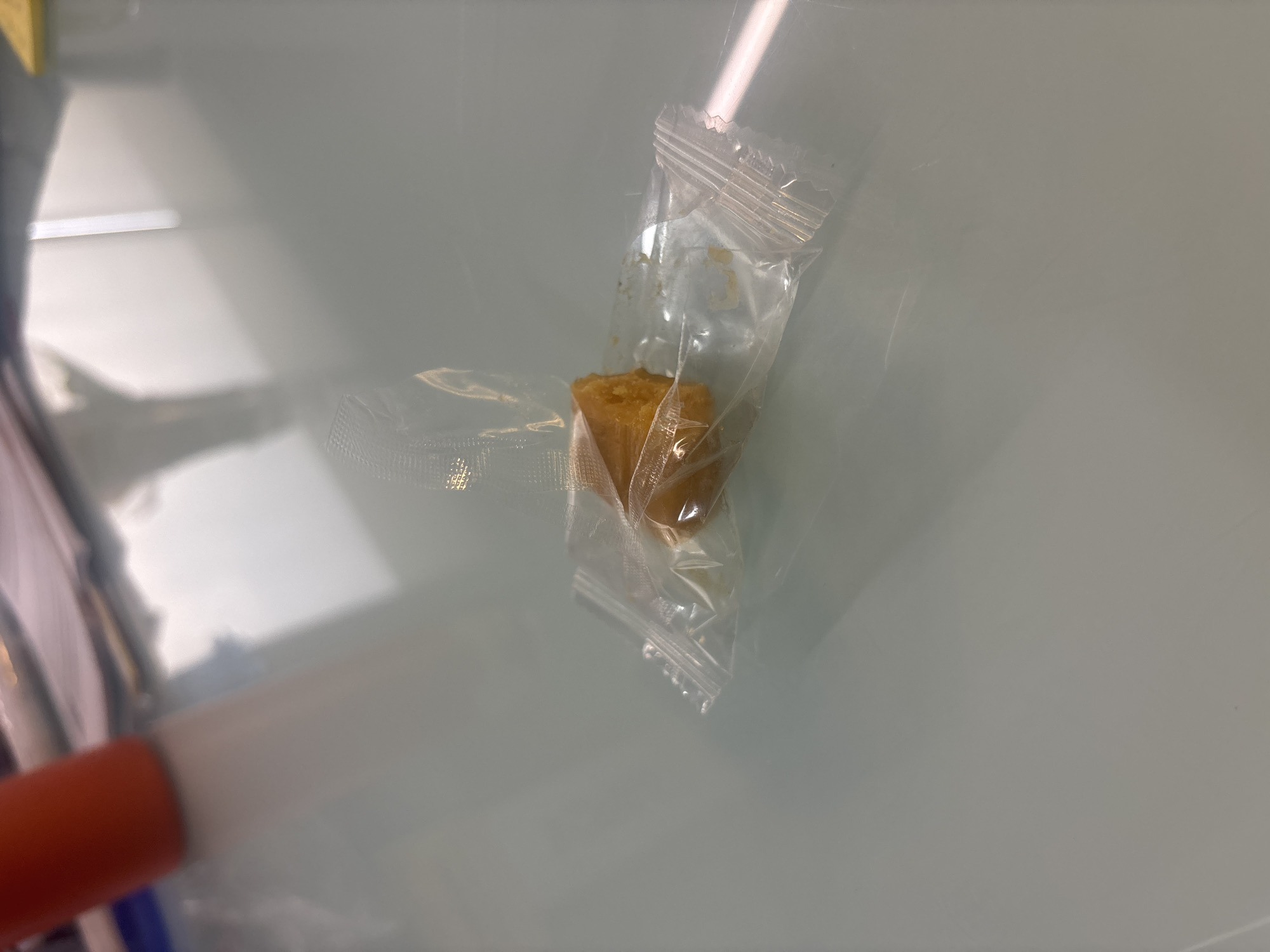 A piece of what appears to be a brown, cube-shaped food item encased in clear plastic packaging sits on a light-colored, reflective surface. The packaging is sealed at the top and bottom with crimped edges. Part of an orange and white object is visible in the lower left corner of the image. The environment is indistinct. The city of Buenavista, Mexico, is not discernible in the image.