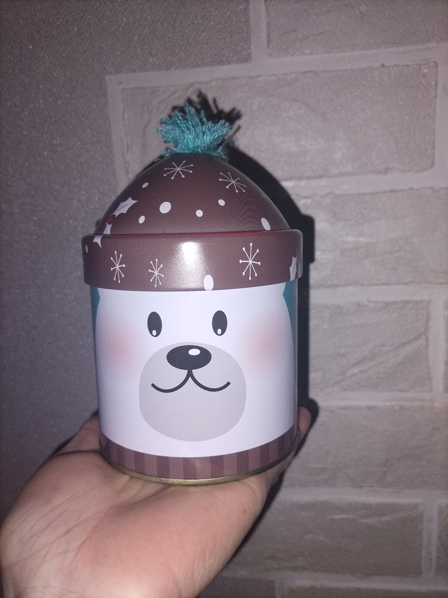 A human hand holds a festive, cylindrical tin container in front of a textured, light brown brick wall. The tin is designed to resemble a cheerful polar bear, with a white body featuring black eyes, a black nose, and pink blushing cheeks. The lid of the container is brown, shaped like a winter hat, and decorated with white polka dots and red and green holly or mistletoe patterns. A fluffy, blue-green pom-pom adorns the top of the lid. Below the lid, a brown band with white snowflake designs encircles the tin, and a striped brown band is visible at the very bottom edge. Shadows from the hand and container are cast on the brick wall, suggesting indoor or low-light conditions. No discernible text is visible on the container.