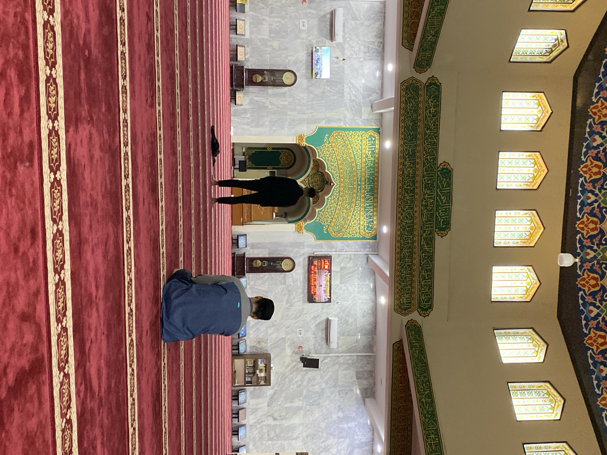 The image shows the interior of a mosque, specifically the Baiturrahman Grand Mosque in Lhokseumawe, Indonesia. The mosque features a large domed ceiling adorned with intricate multicolor patterns. Several tall, arched windows with stained glass are visible along the upper walls, allowing natural light to filter in. The walls are decorated with elaborate green and gold calligraphy. The floor is covered with a plush red carpet marked with parallel lines that denote prayer rows. Two men are present. In the middle ground, one man stands facing away from the camera, dressed in a black long-sleeved shirt and pants. Another man, in the foreground, is seated facing away from the camera, wearing a blue and gray long-sleeved shirt and a dark skullcap. He appears to be in prayer. The overall atmosphere suggests a quiet and contemplative setting.