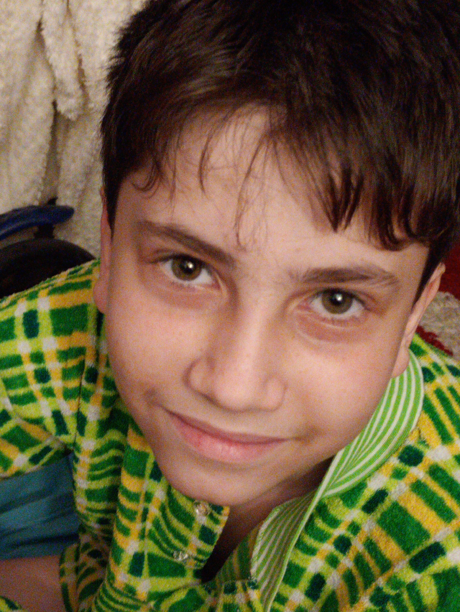 The image is a close-up portrait of a young boy. He has dark hair and brown eyes. He is wearing a green and yellow checkered shirt. The background is out of focus and appears to be a textured blanket. There is no identifiable information within the image to indicate its location in Horishni Plavni, Ukraine.