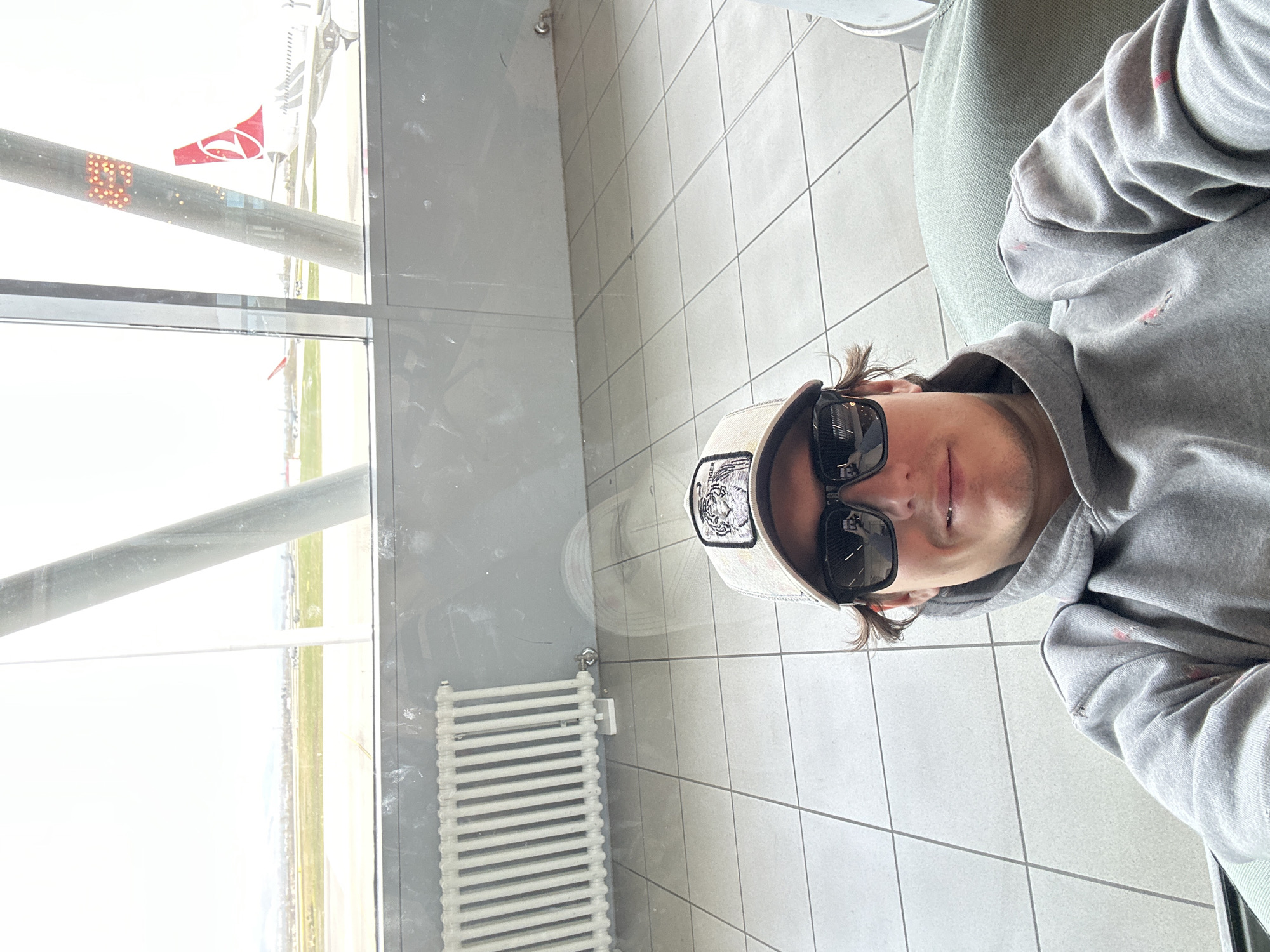 The image depicts a person wearing a grey hoodie and a cap with sunglasses, sitting in what appears to be an airport waiting area. The person is facing the camera. Visible through the window are a plane with the Turkish Airlines logo and part of an airport runway. In the background, there's a radiator. Given the airline and airport runway views, the location is likely the Sofia Airport, near Sofia, Bulgaria. The scene is casual, with the person seemingly waiting for a flight.