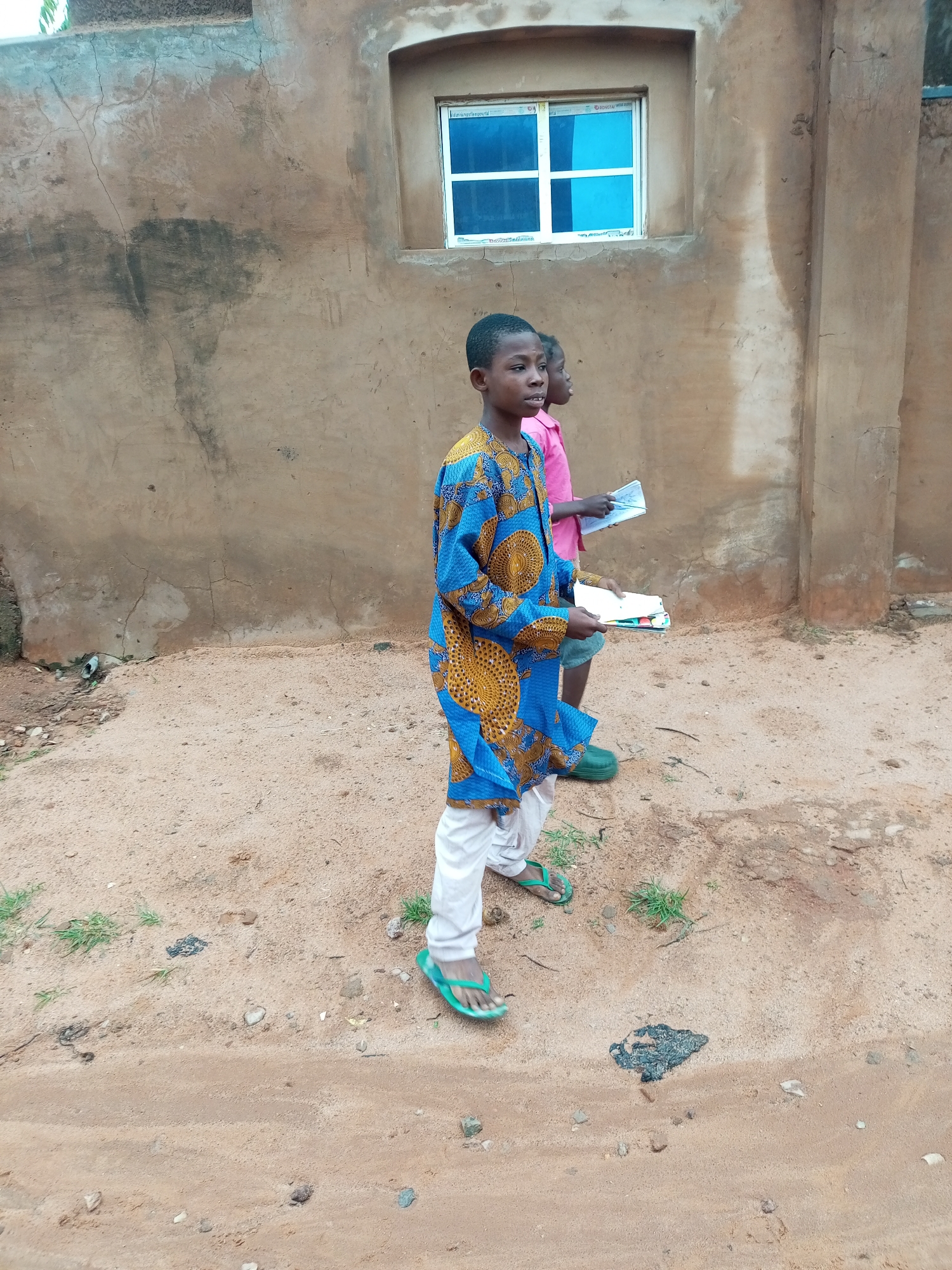 In Tempure, Nigeria, two young boys are walking on a sandy path next to a weathered, ochre-colored building. The boy in the foreground, wearing a vibrant blue and yellow patterned tunic and white pants, carries a stack of colorful notebooks. He is also wearing green flip-flops. Behind him, another boy in a pink shirt holds a book or pamphlet. The building has a small, rectangular window with blue panes. The ground is a mixture of sand and small stones, with sparse patches of green grass.