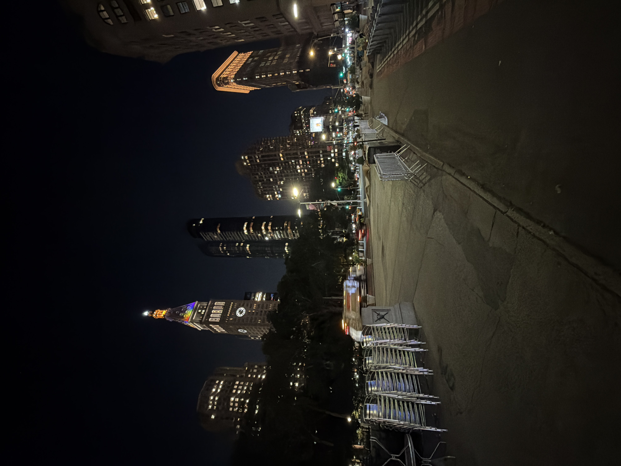 The image captures a night view of Madison Square Park in New York City, with its iconic buildings illuminated against the dark sky. In the foreground, stacks of empty metal chairs are neatly arranged on a paved area, suggesting preparation or closure of an outdoor event. Adjacent to the chairs is a low stone structure.
Beyond the chairs, a street stretches into the distance, with a blurred vehicle in motion, indicating ongoing traffic. Metal barriers line the right side of the scene, separating the walkway from the street. Streetlights cast a warm glow on the path, and trees are visible in the mid-ground, their dark forms contrasting with the lit-up buildings.
The skyline is dominated by several skyscrapers. The most prominent is the Metropolitan Life Insurance Company Tower, also known as the MetLife Tower, its spire adorned with colorful lights, possibly a rainbow. Other notable buildings in the background include the Flatiron Building, identifiable by its distinctive triangular shape and glowing facade, and other high-rise structures with numerous lit windows. The overall atmosphere is that of a bustling urban environment at night.