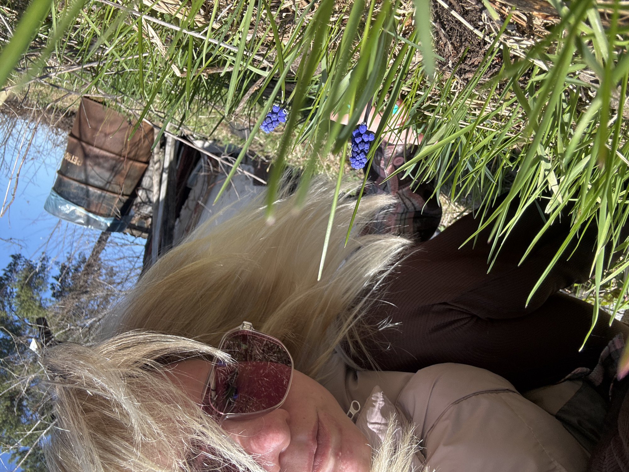 A ground-level, upside-down photograph shows a woman lying among tall, green grass. She wears tinted sunglasses and a beige jacket. Her blond hair spreads around her face. In the background, seen through the grass, are reflections of trees, sky, and a metal barrel, suggesting a watery surface. Two clusters of blue muscari flowers are also visible in the foreground. The location is Chaikovsky, Russia.