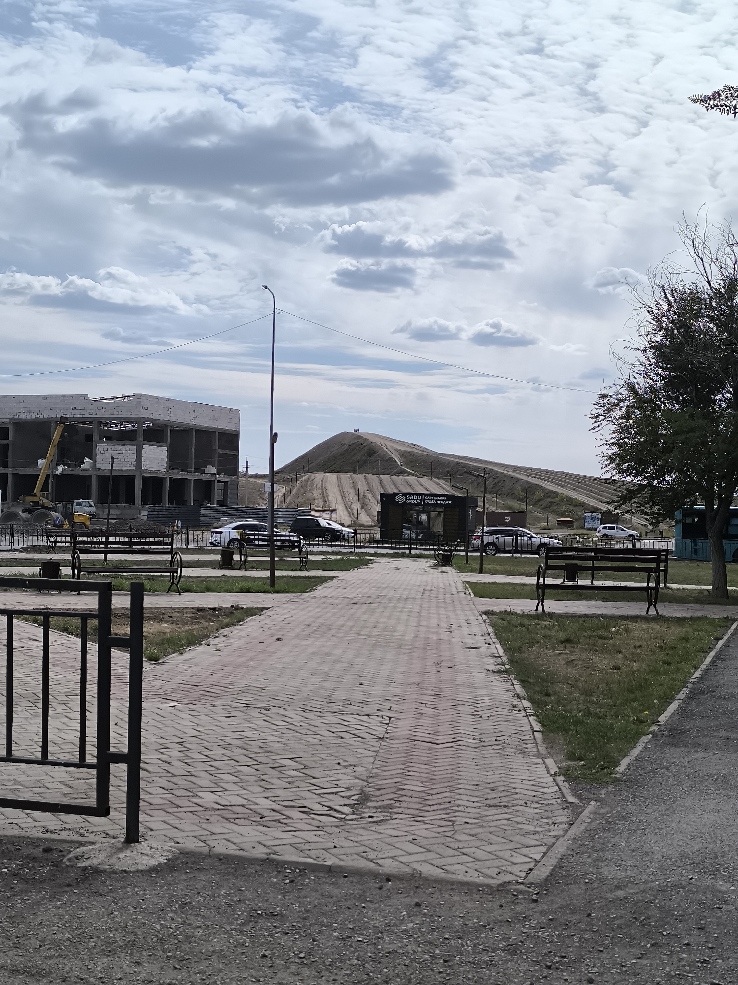 The image depicts a construction site in Qarağandı, Kazakhstan. In the foreground, a paved walkway made of red bricks leads into the scene. To the left, a black metal fence borders the walkway. In the mid-ground, several benches are visible, and a construction site with a multi-story building under construction is prominent. A yellow crane and other construction equipment are present. Cars are parked to the right of the construction site. In the background, a large mound of earth dominates the landscape, with some dirt roads visible on its slopes. The sky is cloudy, with a mix of white and gray clouds. A street lamp stands near the center of the image, and power lines are visible stretching across the sky. A tree with dark foliage is located on the right side of the frame. A small building with signage that reads "SADU | САТ БІЛІМ GROUP | ОТДЕЛА ПРОДАЖ" is also visible, positioned in front of the mound. The overall scene suggests an ongoing development or land alteration project within an urban or semi-urban environment.