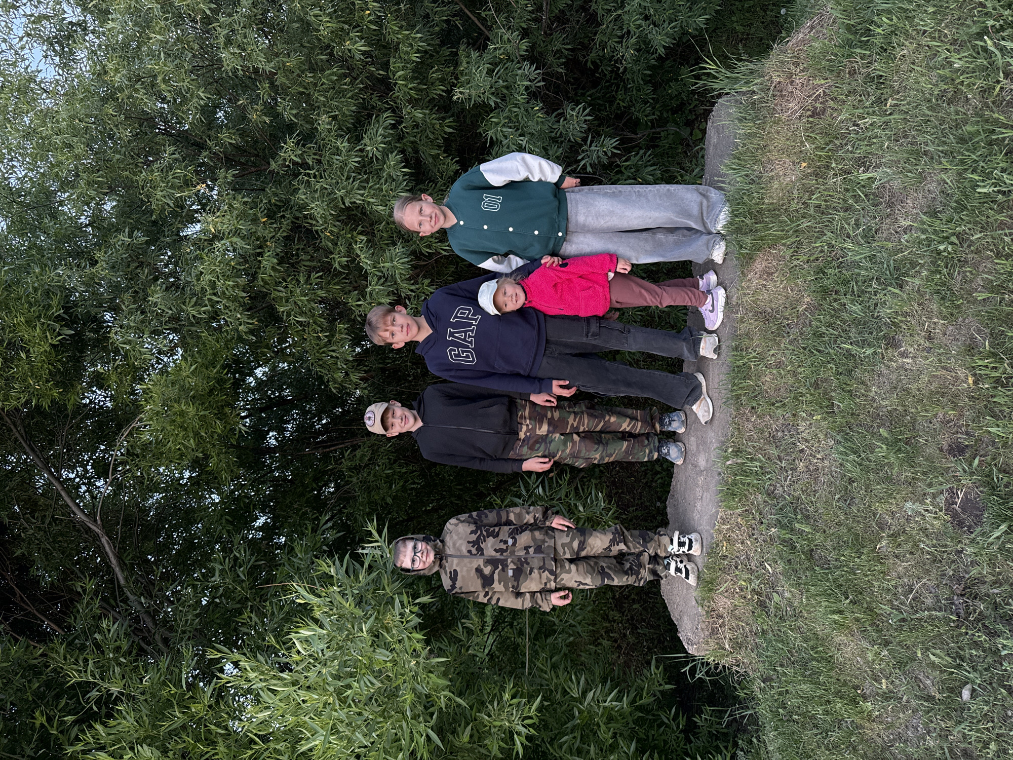 Four children stand on a concrete ledge bordered by grass and trees. The tallest child, a girl, wears a green and white varsity jacket and grey sweatpants. Next to her is a boy in a navy blue GAP sweatshirt and a younger child in a pink jacket and white hat. The two are positioned close together. Below them is a boy in a black zip-up jacket and camouflage pants, topped with a baseball cap. The last child, at the bottom, wears glasses and a full camouflage outfit. The lush trees form a backdrop to the group. No specific location in Горный, Russia can be determined based solely on the provided image.