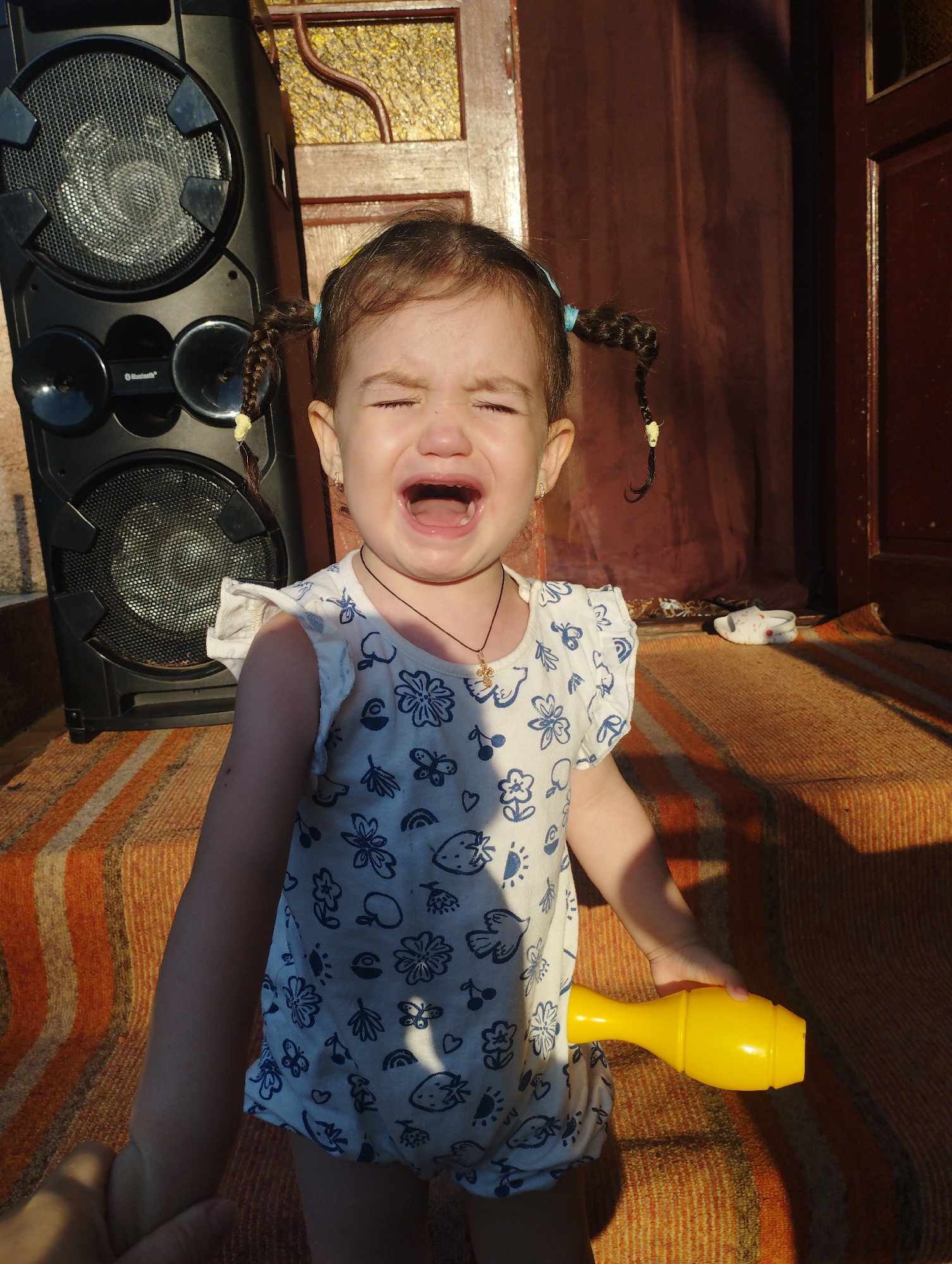 A young girl with pigtails is crying loudly, with her eyes closed and mouth open wide. She is wearing a white dress with a blue floral pattern and a gold cross necklace. She is holding a yellow toy in her right hand. The girl is standing on a striped rug in front of a large black speaker. In the background, there is a brown curtain and a wooden door. The scene appears to be outdoors, possibly on a porch or patio, with sunlight casting shadows. This image was taken in Fălești, Moldova.