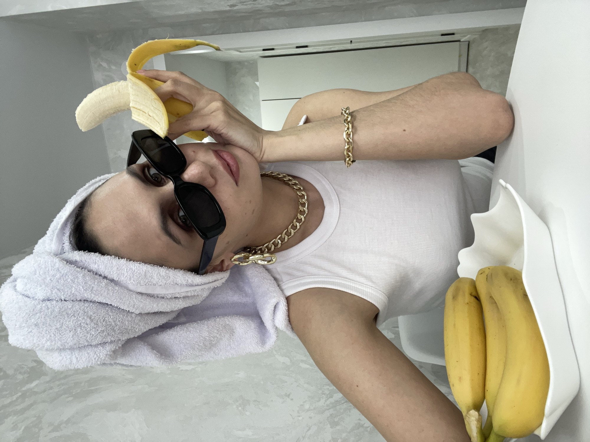 A woman with fair skin is shown from a slightly skewed, close-up perspective. She is wearing a white ribbed t-shirt and has a white towel wrapped around her head. Black sunglasses are perched on her face. She is wearing a gold chain necklace, a gold bracelet, and gold earrings. The woman is holding a partially peeled banana near her head. A white container holding two unpeeled bananas is visible to the right of the frame. The background appears to be a white or light gray wall and counter. The location is not discernible from the image; there is no indication it is in Izhevsk, Russia.