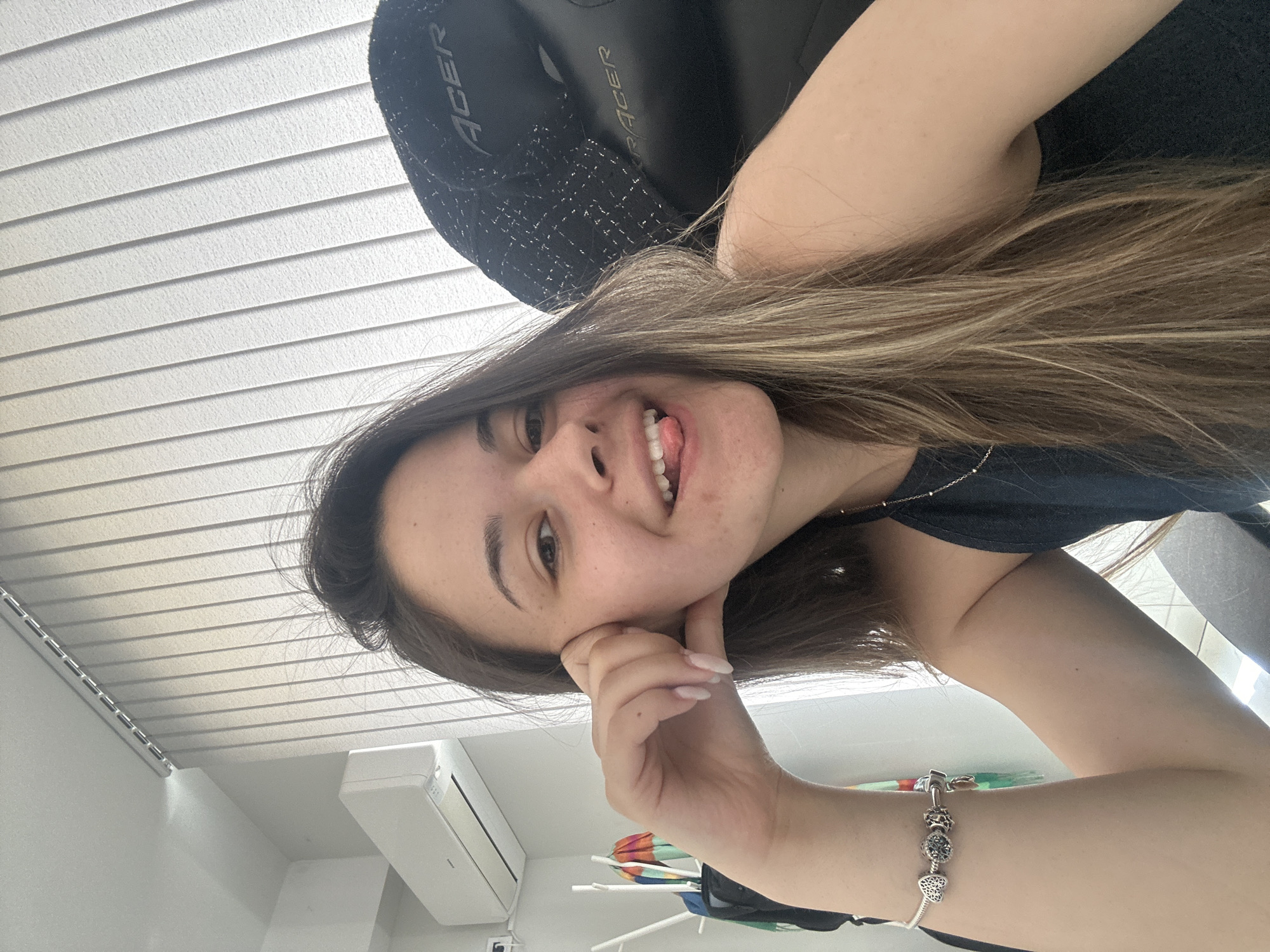 A young woman with long brown hair is smiling and sticking her tongue out in a selfie. She is wearing a bracelet on her left wrist. She is sitting in a black Acer gaming chair. The background includes white window blinds, a white air conditioning unit, and a clothes drying rack with colorful items. There is no information available to determine that the location of the photo is in Sofia, Bulgaria.