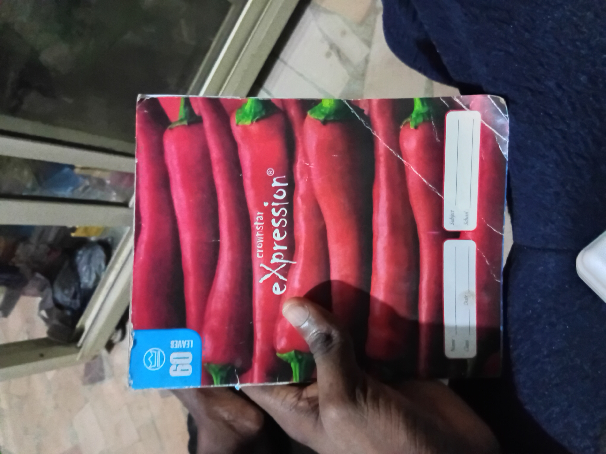 The image shows a notebook with a red chili pepper design on the cover, held by a person's hand. The notebook has "60 LEAVES" printed on a blue tab at the top left. The brand name "crownstar Expression" is visible in white text on the cover. There are also labels for "Name", "Date", "Class", "Subject", and "School" at the bottom of the notebook, with blank lines for writing. The background suggests an indoor setting with a tiled floor and a piece of furniture, possibly a cabinet or table, with a dark object inside. The lighting appears to be artificial. The image is cropped and slightly angled, focusing on the notebook and the hand holding it. The scene is static and does not depict any particular event or action.