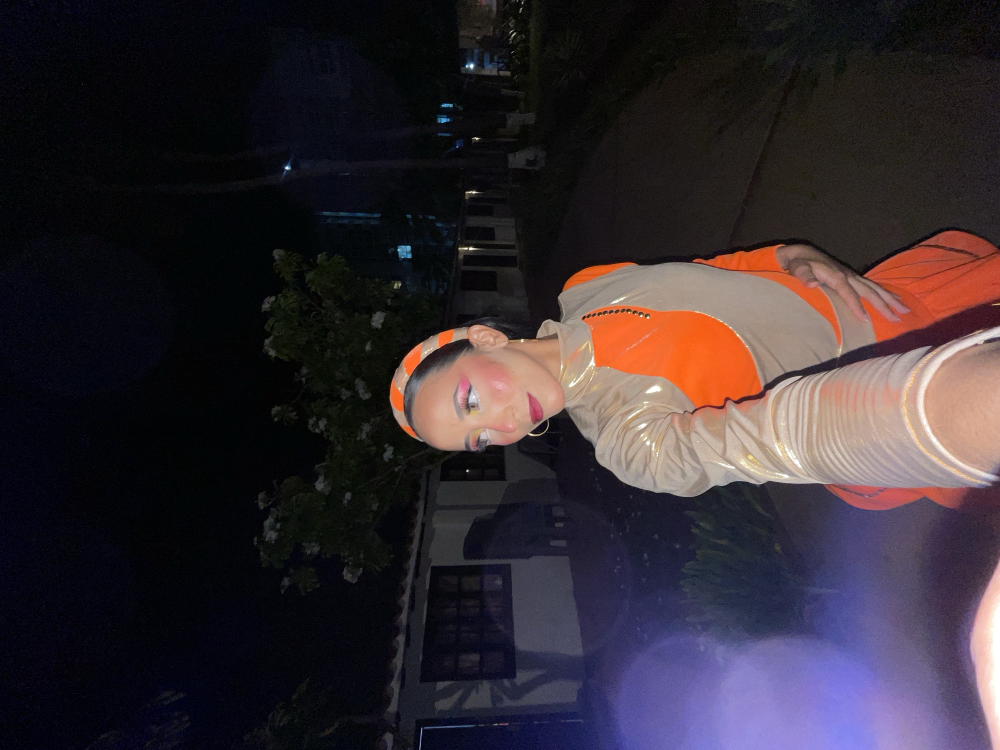 A person wearing an orange and tan outfit with a matching headband poses for a selfie at night. The person has bold makeup, including pink eyeshadow and red lipstick. They are standing on a paved path next to a building with windows and a tiled roof. Trees and foliage are visible in the background, and there is a slight lens flare in the foreground. The scene appears to be outdoors, possibly in the city of Porlamar, Venezuela, given the architecture and vegetation.