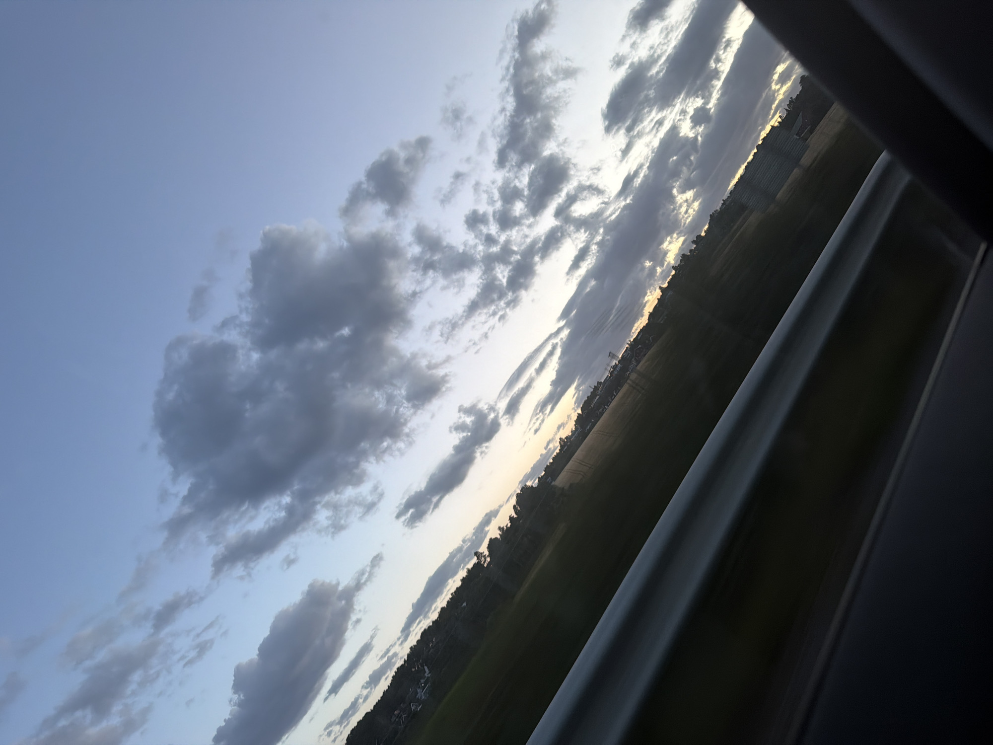 The image depicts a view from inside a vehicle, looking out at a landscape during what appears to be dusk or dawn. The horizon is tilted, suggesting the vehicle is in motion or the photo was taken at an angle.
In the foreground, a dark, metallic railing runs diagonally across the lower left of the frame, likely part of the vehicle's interior or a barrier seen through a window. Below this railing is a dark, out-of-focus area, possibly the dashboard or interior of the vehicle.
The middle ground features a stretch of green fields, with a few trees and some distant buildings or structures visible along the tree line. The fields show some texture, possibly indicating cultivation or tire tracks. The sky is a gradient of pale blue to a lighter hue near the horizon.
Scattered across the sky are numerous clouds, varying in size and density. Some are dark and appear to be storm clouds, while others are wispy and lighter, allowing the sky's color to show through. The lighting suggests a low sun position, casting a soft glow on the horizon.
While the image is oriented to show a landscape, no specific landmarks or features definitively place it within Sarpsborg, Norway. The environment appears to be a rural or semi-rural area, possibly on the outskirts of a town or city. There are no people or discernible actions taking place in the image.