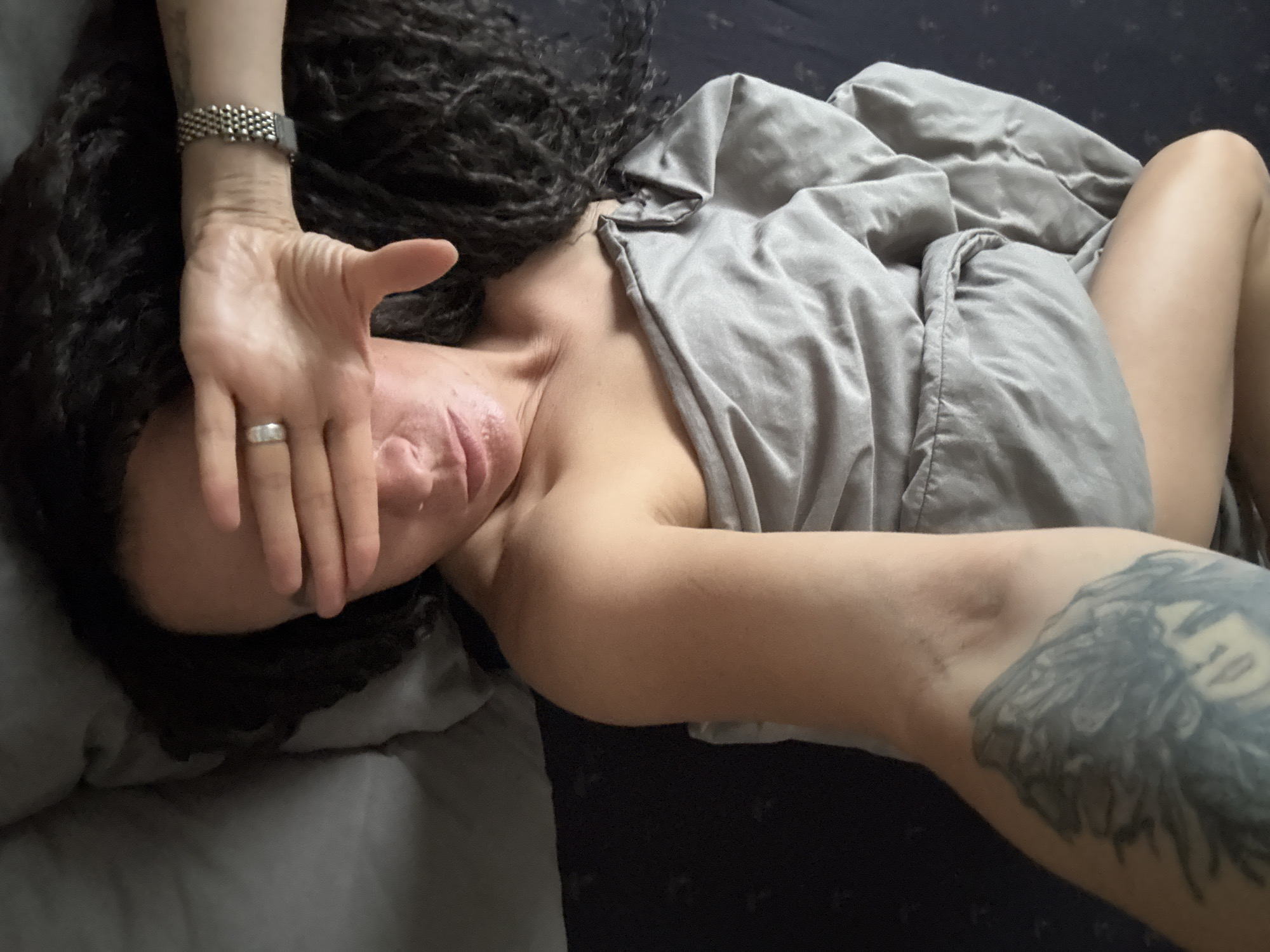 An overhead shot captures a person lying on their back in bed, partly covered by a light gray blanket. The individual has long, dark, curly or dreadlocked hair that spreads across dark-colored bedding. Their left hand is raised, covering their face, with a simple silver-colored band ring visible on the ring finger. A silver-toned watch with a beaded or mesh strap is worn on the left wrist. The right arm is extended outwards, revealing a large tattoo on the upper arm, depicting what appears to be a face, possibly female, framed by feathers or wings. A portion of the person's right leg or thigh is visible from under the blanket in the lower right of the frame. The scene is set indoors, likely in a bedroom. The person's face is obscured by their hand, making their expression and emotions unreadable; the pose could suggest resting, shielding from light, or privacy. There is no visible text in the image. The location context provided is Chaikovsky, Russia.