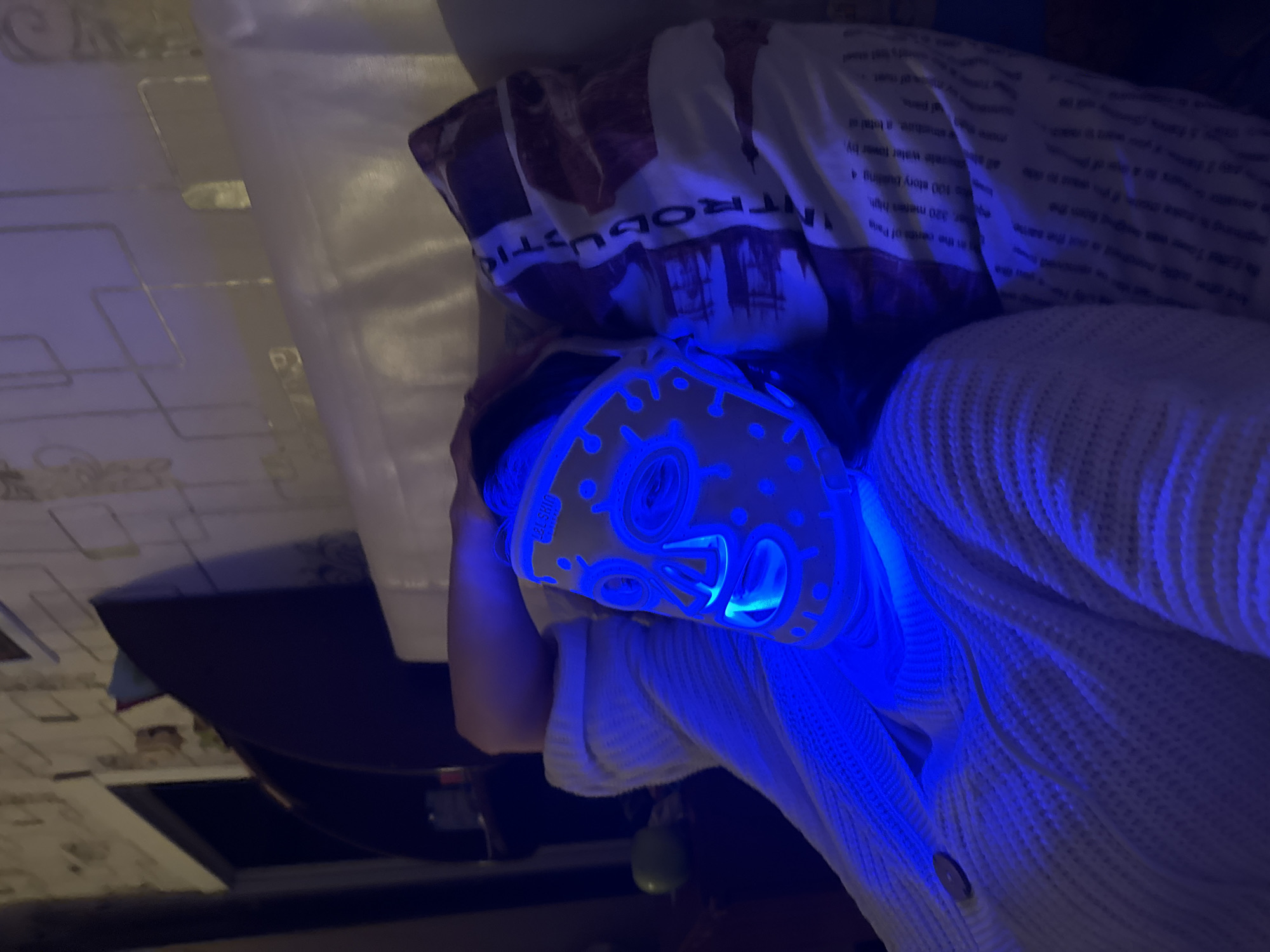 A person is lying in bed wearing a light-up blue mask. The bed has a pillow with text that says "INTRODUCTION" on it. The person is wearing a white sweater. The scene is indoors and dimly lit. The location in the city is unknown.