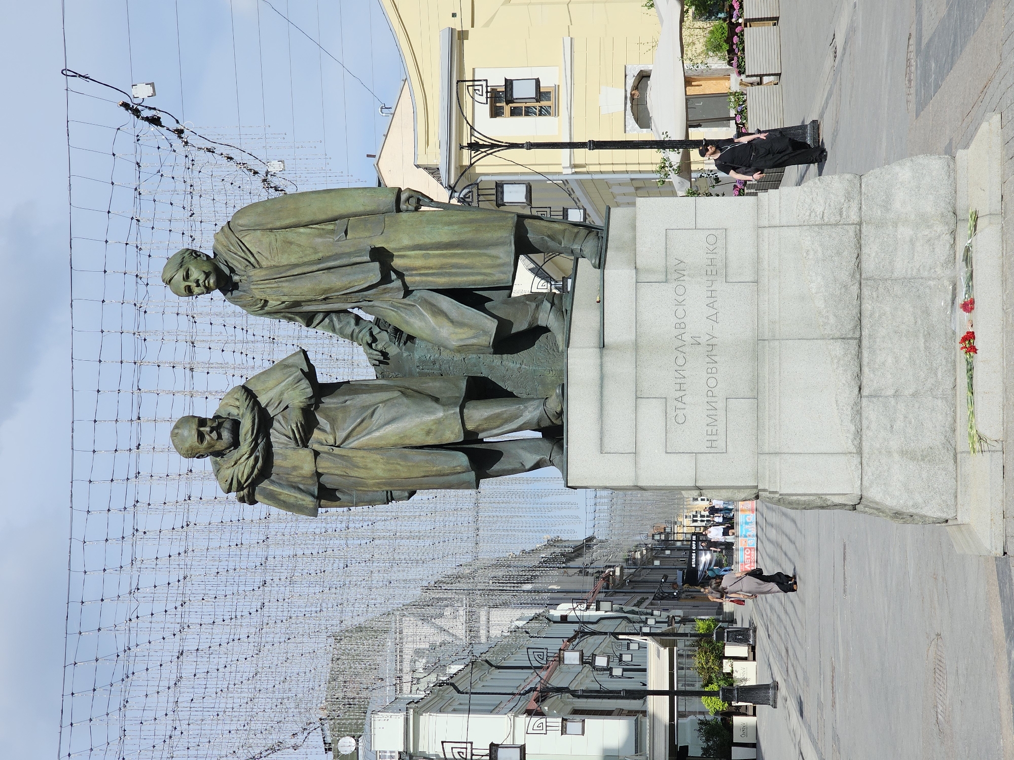 The image shows the monument to Stanislavsky and Nemirovich-Danchenko in Moscow, Russia. The bronze statue features two men in overcoats on a stone pedestal. Text in Cyrillic script is engraved on the pedestal. A grid of string lights hangs overhead. Two people are near the monument. One is facing the pedestal wearing all black and the other is further away walking on the sidewalk. Several flowers lay at the base of the monument. Buildings line the street.