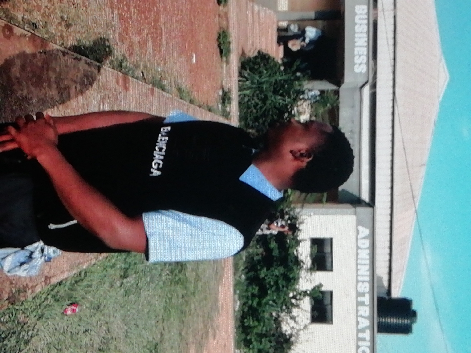 A young man stands outside a building that appears to be an academic institution. The building has sections labeled "Business" and "Administration". The location is Enugu, Nigeria, as indicated by the uniform worn by the man, which aligns with school uniforms commonly seen in that region. The man wears a blue collared shirt and a black vest with "Balenciaga" printed on it. He has his arms crossed in front of him and is looking upwards. The environment includes grass and a concrete walkway near the building. A red object is on the grass.
