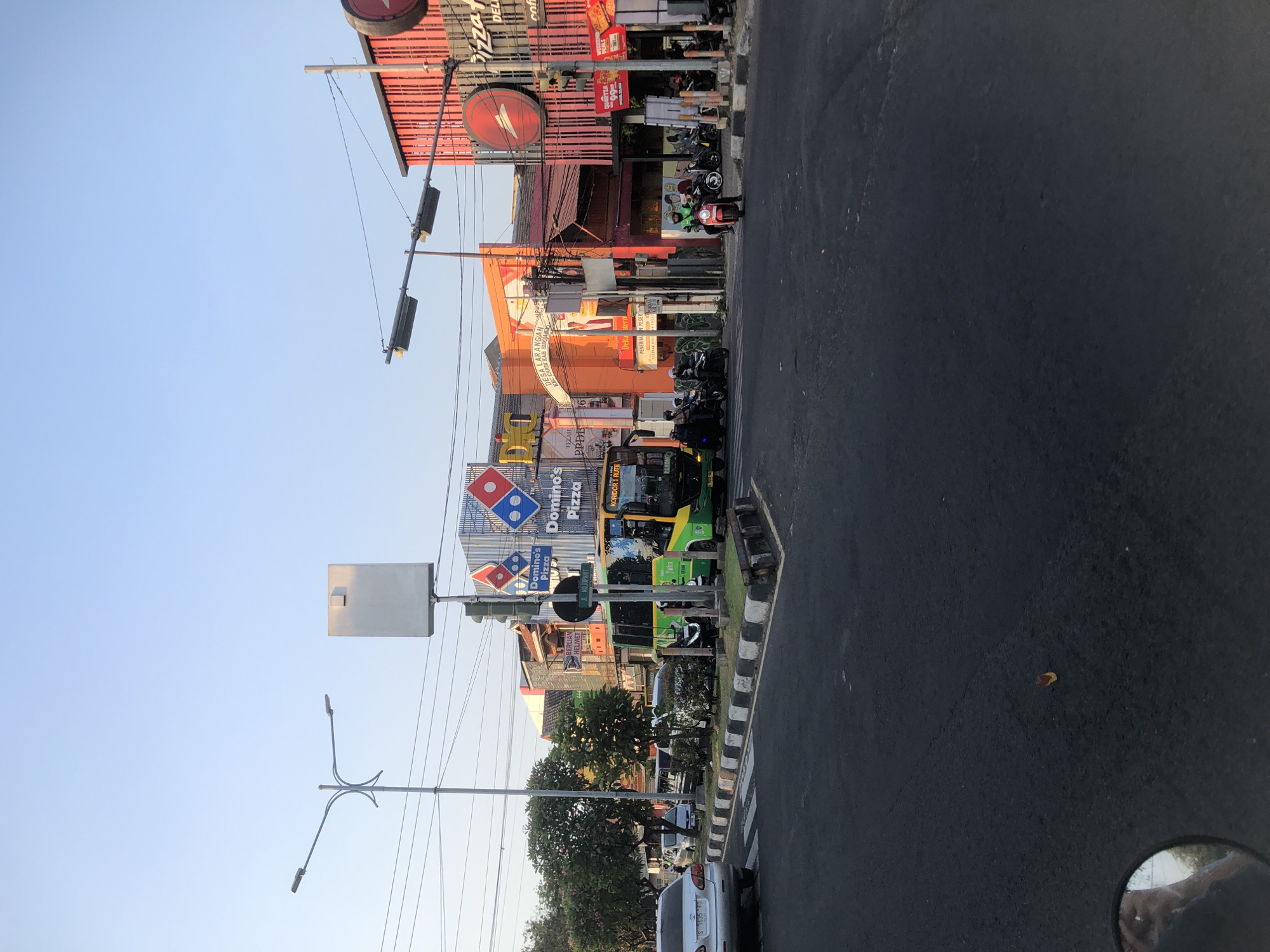 The image depicts a street scene in Larangan, Indonesia, likely at an intersection during daylight hours. A green and yellow bus, marked with "KORIDOR 1 RUTE" and "CEBAN", is stopped at the intersection, its doors open and passengers visible. Several motorcycles with riders are also present, some waiting at the intersection, others in motion. Buildings line the street, including prominent signage for "Domino's Pizza" and "Pizza Hut". A traffic light is visible, as is a street light. The road is paved, with clear lane markings and a pedestrian crossing. The sky is a clear blue, suggesting good weather. The overall impression is that of a typical urban street in Indonesia, with active transportation and commercial establishments.
