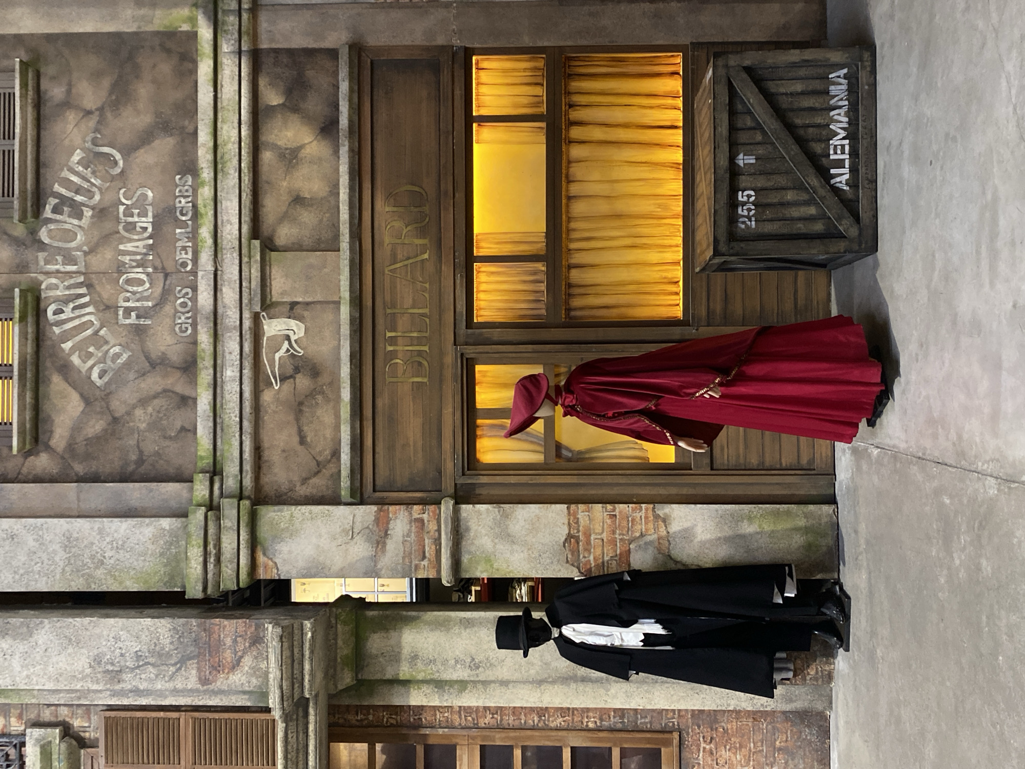 The image presents a detailed diorama or set, resembling a narrow European street scene, likely situated indoors. Two life-sized figures, appearing to be mannequins, are centrally placed on a plain concrete floor.
On the right side of the frame, a figure dressed in a vibrant red cloak and matching red hat stands beside a shop entrance. The shop features a wooden facade with "BILLARD" in gold lettering above warmly illuminated windows, which are covered by yellow curtains, suggesting an evening setting. Adjacent to this shop, a wooden crate is visible, stenciled with "255", an upward arrow, and the word "ALEMANIA" (Germany).
Further left, a second figure is dressed in a classic black suit, white shirt, and a black top hat. The surrounding buildings exhibit textured facades with imitation stone and brickwork, displaying weathered effects and subtle traces of moss or grime. Another shop sign is visible on the left, bearing the French words "BEURRE OEUFS FROMAGES" (Butter, Eggs, Cheeses) and "GROS OEMLGRBS", accompanied by a white silhouette of an animal, possibly a cow or goat.
The scene is static, depicting a snapshot of a bygone era. The blend of French shop names and the German crate label strongly suggest a Western European influence. Although the scene is set in Europe, the context indicates the photograph was taken in Buenos Aires, Argentina, implying this is an exhibition or themed area.