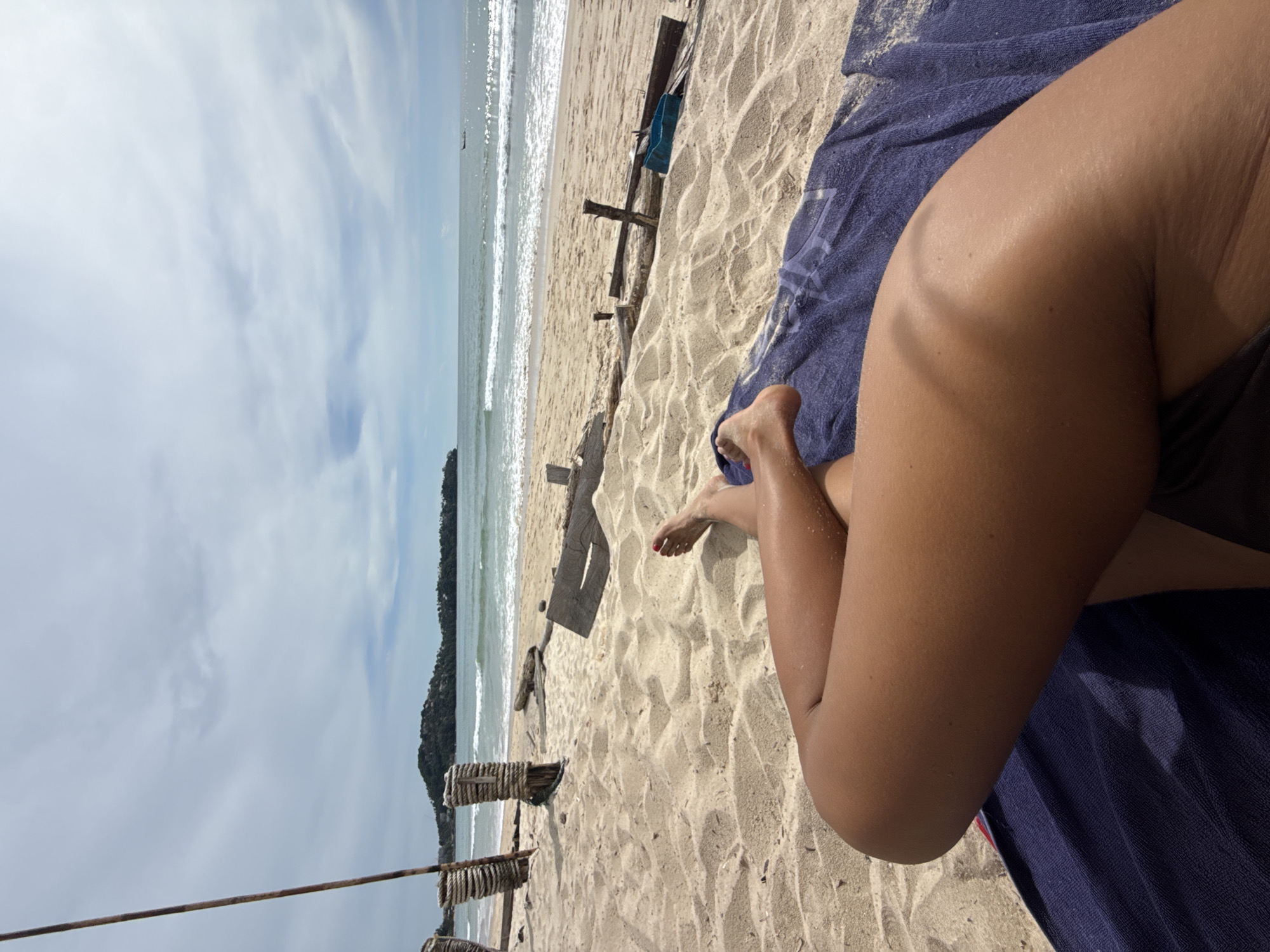 The image shows a low-angle shot of a person's legs and feet relaxing on a sandy beach in Choeng Thale, Thailand. The person is lying on a blue towel, with their legs crossed. Their feet are visible, with red nail polish on one foot. The beach is covered in light-colored sand with some debris, including wooden posts and planks, scattered around. In the background, the turquoise ocean meets a cloudy sky, with a small boat visible on the horizon. A hilly, tree-covered coastline is also visible in the distance. The overall scene depicts a tranquil beach setting.