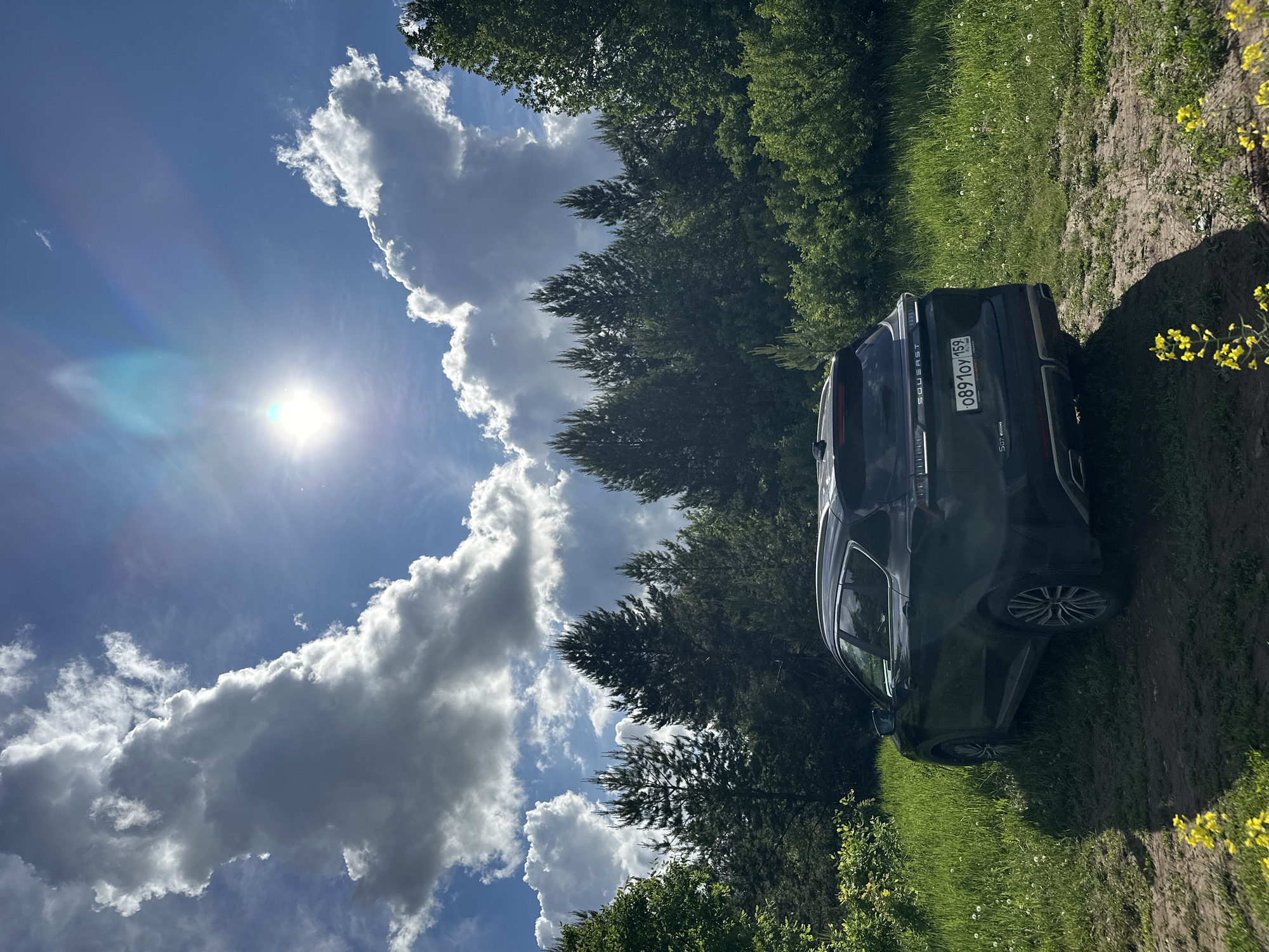 The image shows a dark-colored SUV parked on a grassy, dirt path next to a field of green grass. The background consists of a forest with tall trees. Above the trees, a blue sky is visible with large, white clouds and a bright sun. The license plate of the SUV is partially visible, showing "0891OY 159." The scene suggests a rural or wooded area. There is no evidence to confirm that the location is the city of Sarapul, Russia.