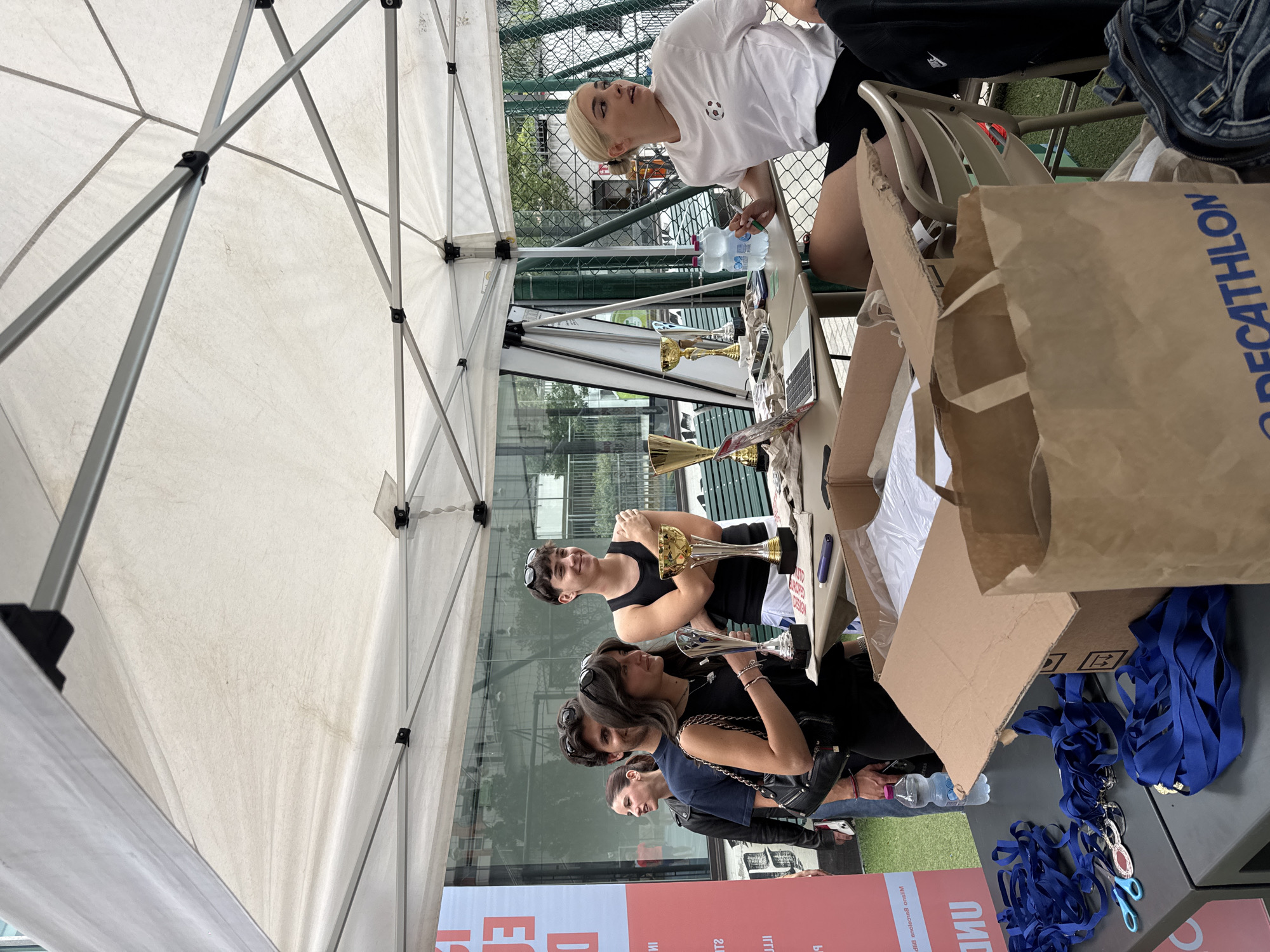 An outdoor scene in Milan, Italy, captures an event under a white pop-up canopy tent, likely at a university sports facility, given the discernible Italian text on banners and water bottles. The ground is covered with green artificial turf.
In the midground, a light-colored rectangular table serves as a reception or awards desk. On the left, four young adults stand in a line. The foremost is a woman with dark hair and sunglasses on her head, wearing a black top and carrying a black shoulder bag, holding a silver trophy and smiling towards the right. Behind her, a man with dark hair and sunglasses on his head wears a dark blue t-shirt and smiles in the same direction. Further back, a woman in a black leather jacket smiles towards the left, with another person partially visible behind her. On the right side of the table, a blonde woman with her hair in a bun, wearing a white t-shirt with a small, colorful soccer ball emblem and black shorts, sits facing right, holding a green marker pen.
The table holds three trophies: two gold-colored and one silver-colored. An open silver laptop, two clear plastic bottles of "Acqua Minerale Naturale," a purple pen, and various small papers or cards are also present. In the foreground on a lower table surface, there are numerous coiled blue ribbons, some attached to circular red and gold medals, and a pair of small blue scissors. An open brown cardboard box, likely containing white packaged items such as t-shirts, is positioned prominently. A large brown paper bag with the "DECATHLON" logo is partially visible in the bottom right corner, next to a denim bag.
In the background, a red and white banner with partially inverted Italian text ("DI E" and "EU" visible, suggesting a university event, possibly "Università degli Studi di Milano") stands behind the left group. Behind the table and further back, a green wire mesh fence separates the event area from a sports court. Trees and modern glass-fronted buildings of an urban environment are visible beyond the fence.