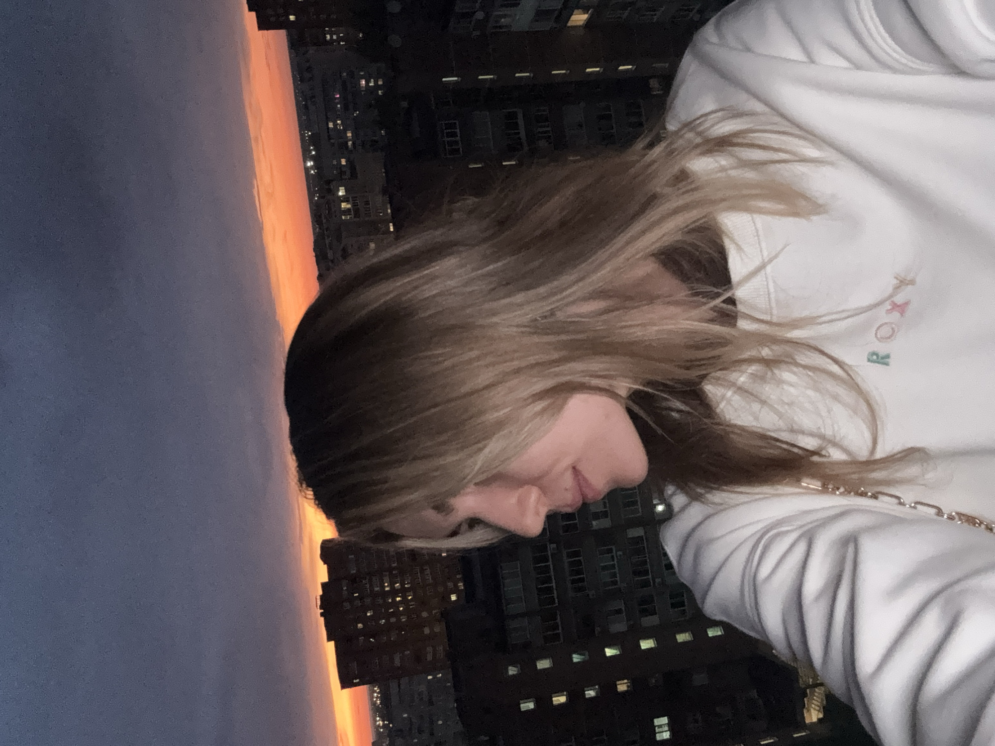 A young female, with blonde hair styled with a side part, is depicted in a profile selfie looking towards the left. She wears a white crew-neck sweatshirt featuring a pastel-colored embroidered "ROXY" logo on the chest. A gold-toned chain, likely from a bag strap, is visible on her right side. The scene is captured at dusk or sunset from an elevated perspective overlooking an urban environment in Unknown, Unknown. The sky transitions from a dark purplish-blue at the top to a vibrant orange-red band stretching across the horizon. Numerous multi-story buildings, largely silhouetted against the sky, occupy the mid-ground and background, with many windows displaying interior illumination.