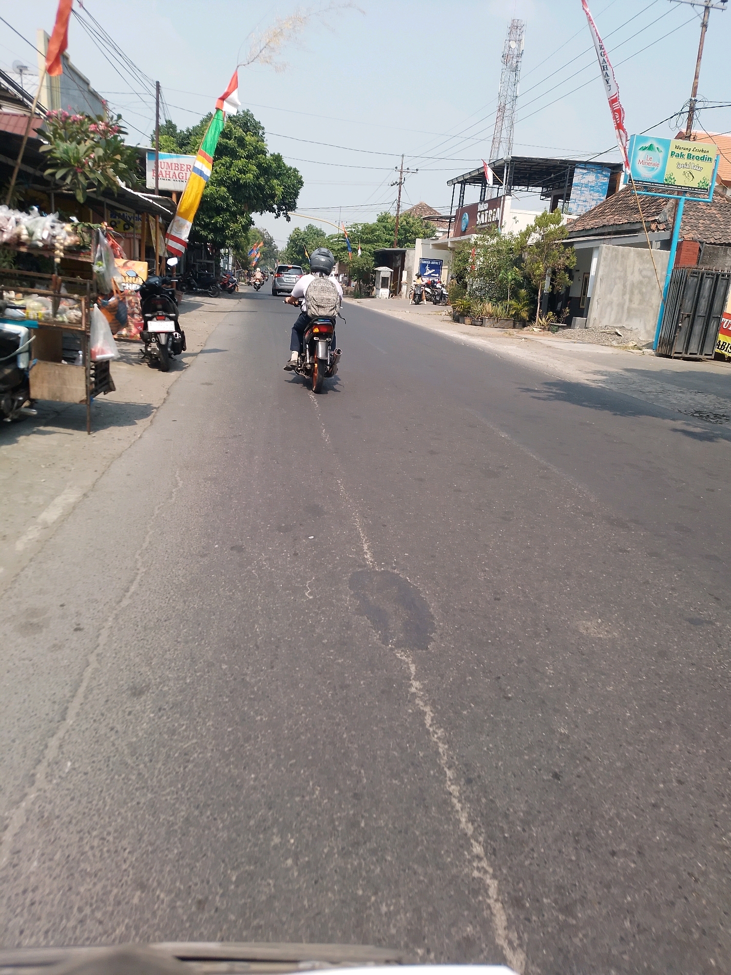 A motorcycle rider wearing a helmet and a backpack is seen from behind, traveling on a two-lane asphalt road. The rider is on the right side of the road, and there is an oncoming car in the distance. On the left side of the road, there are several shops and stalls, including a stall selling food or produce. Indonesian flags and banners are visible, indicating a local event or celebration. The environment appears to be a typical street scene in a town or city, possibly in Indonesia, given the context of Pandan Toyo. The buildings on the right side of the road are a mix of residential and commercial structures. A telecommunications tower is visible in the background. The weather appears to be clear and sunny.