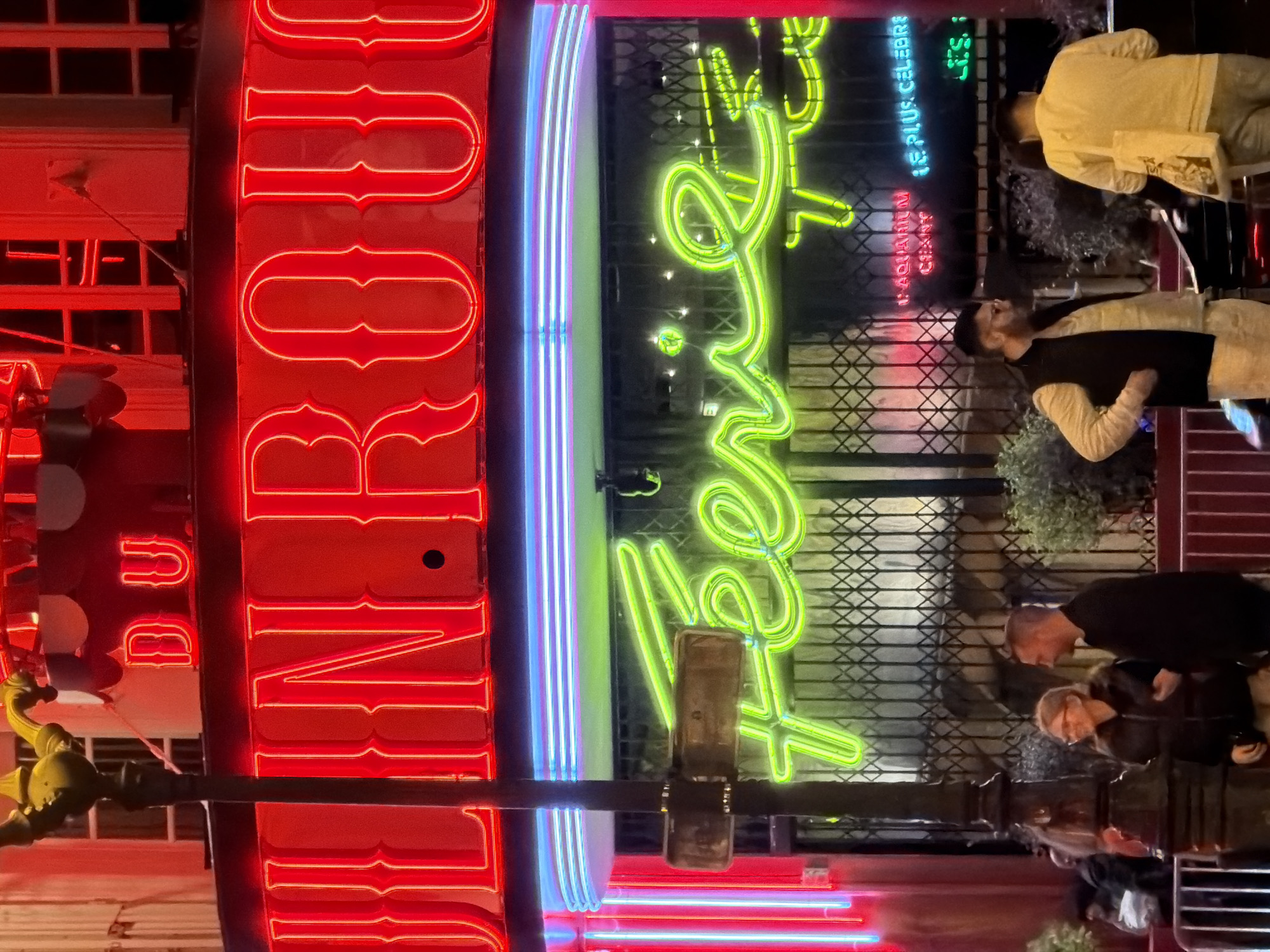 The image shows a street scene in Paris, France, at night. The primary focus is on two neon signs, one red and one green, mounted above a storefront or establishment. The red sign displays "Rendez-Vous" vertically in bold, illuminated letters. Adjacent to it is a sign with vertical blue and white neon stripes. The green sign, also in neon, has a cursive design and includes text such as "L'AQUARIUM CHAOS" and "LE PLUS CÉLÉBRÉ". A group of people are standing in front of the signs, appearing to be pedestrians or possibly patrons of the establishment. They seem to be talking and interacting. There is a decorative metal gate behind the green neon sign.