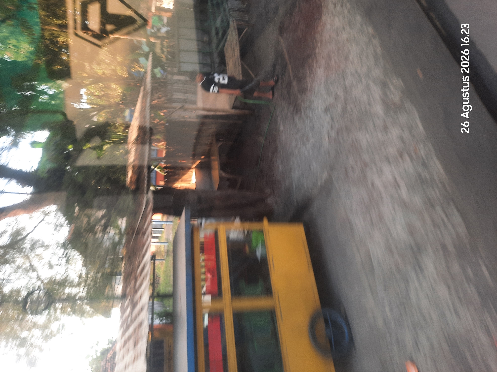In Sukokerto, Indonesia, on August 26, 2026, at 4:23 PM, a yellow food cart with red and green compartments is positioned on the left side of a dirt road. A young person wearing a black and white jersey with the number 26 on it is seen on the right side of the road, holding a green hose. The background includes trees, a rustic structure with a corrugated tin roof, and wooden tables. The scene appears to be outdoors, with natural light filtering through the trees, suggesting a sunny day. The overall image has a blurred quality, possibly due to motion.