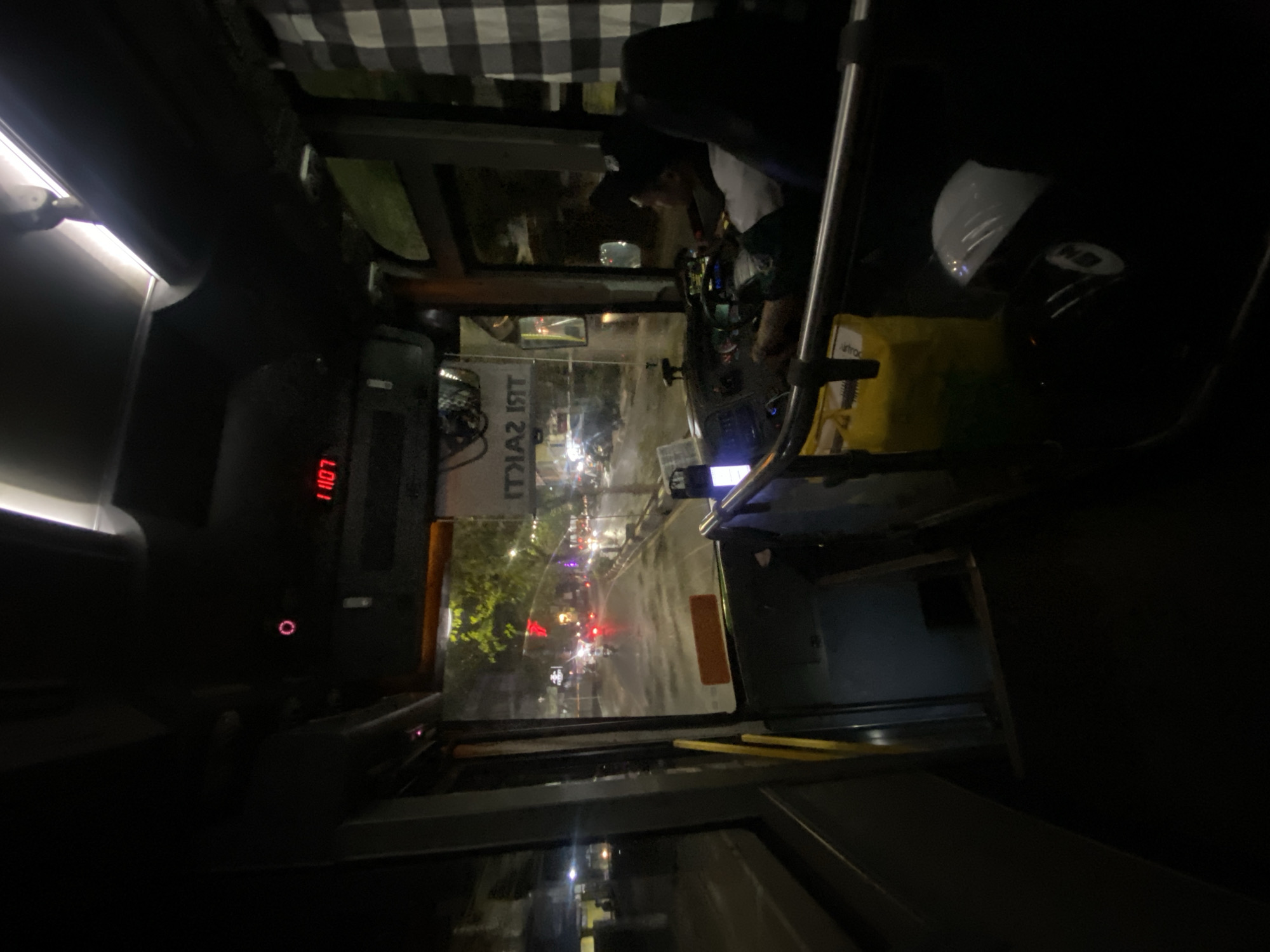 The image shows the interior of a bus at night, likely in Banjarmasin, Indonesia, with a view of the road ahead through the windshield. A driver, wearing a dark shirt and a cap, is seated at the steering wheel, looking forward. The bus is illuminated by the dashboard lights, a red digital display showing "1107", and the streetlights and vehicle lights from outside. The road appears wet, suggesting it may be raining or has recently rained. Trees and buildings are visible along the roadside. A checkered curtain hangs on the right side of the bus. A yellow bag and a white helmet are visible in the foreground, on the right side of the bus. The overall lighting is dim, creating a dark atmosphere typical of nighttime urban scenes.