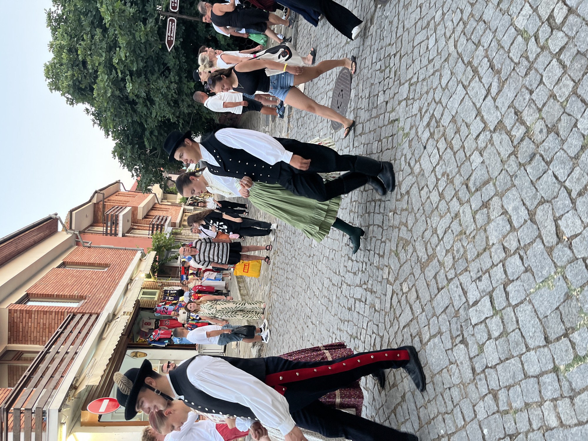 In Nesebar, Bulgaria, a group of people are walking on a cobblestone street lined with shops and brick buildings. In the foreground, two individuals in traditional folk costumes are walking. The person on the left wears a black hat adorned with feathers, a white shirt, a black vest, and black trousers with red detailing. The person on the right wears a similar black vest and hat, a white shirt, and a green pleated skirt. Behind them, other people are walking, some in casual clothing, while others are browsing in the shops. The atmosphere suggests a cultural event or a traditional celebration.