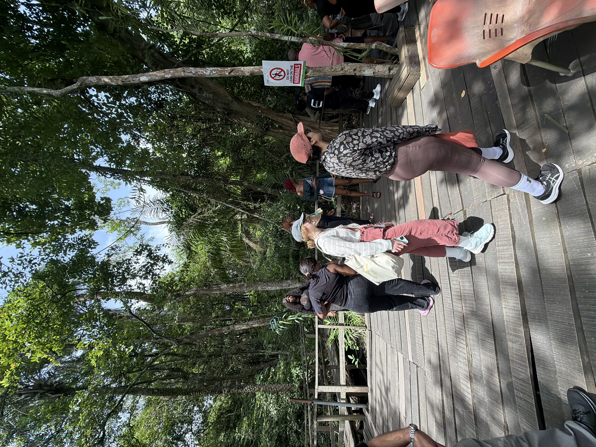 The image shows a wooden walkway surrounded by dense green trees, likely located in a forested or park-like environment. A group of people is standing on the walkway, some appearing to observe the surrounding scenery. There is a sign that reads "DO NOT LEAN ON THE HANDRAILS." The general location is the city of Lekki, Nigeria. The walkway seems to be elevated, with a railing visible along its side. The lighting suggests it is daytime.