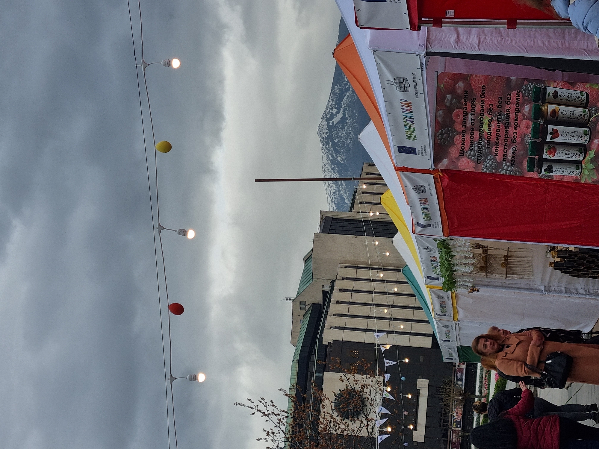 The image shows an outdoor scene in Sofia, Bulgaria, likely at a farmers market (indicated by the Cyrillic text "ФЕРМЕРСКИ ПАЗАР" meaning "Farmers Market"). Stalls are set up with produce and goods for sale. The stalls have colorful awnings in red, orange, yellow, and white. A woman is standing in the foreground wearing a beige coat and carrying a black purse. There are other people nearby, including individuals wearing dark jackets and a red jacket.
Overhead, a string of lights hangs with colorful ball decorations in red and yellow. There's also a row of white triangular pennant flags strung along another line.
In the background, a modern-style building is visible. Beyond the building, there is a view of a mountain range, with snow visible on the peaks. The sky is overcast with grey clouds.