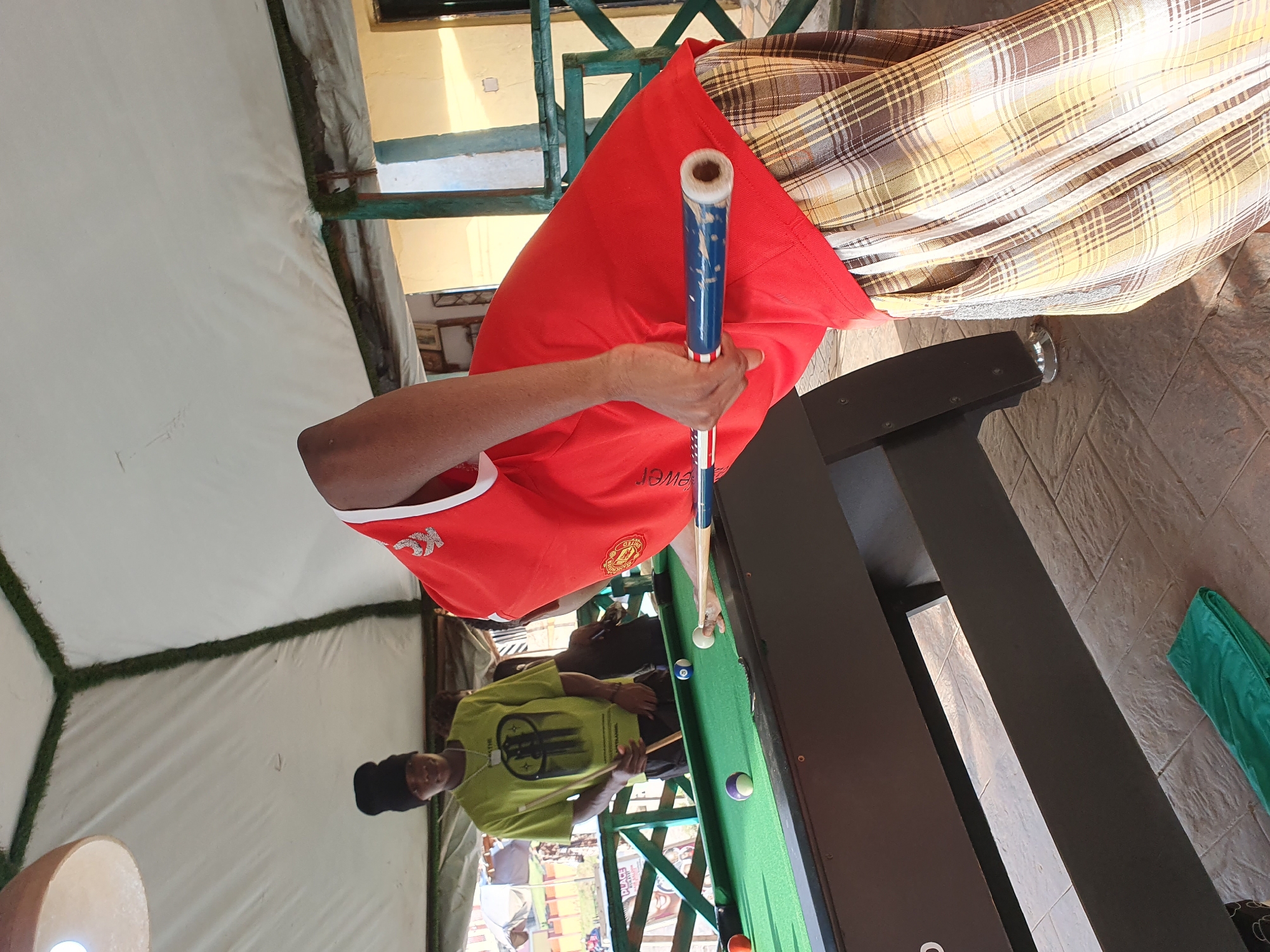 In Lokoja, Nigeria, two individuals are engaged in a game of pool. One person, wearing a black beanie and a green t-shirt, stands holding a pool cue and looking towards the pool table. The other person, prominently featured in the foreground, is bent over the pool table, in the process of striking a billiard ball with a cue. This person is wearing a red shirt and a plaid skirt or wrap. The pool table is green, with several colored balls visible. The setting appears to be an outdoor or semi-outdoor area, possibly a recreational space, with a white tent-like covering overhead and wooden railings. The ground is paved with stone tiles.