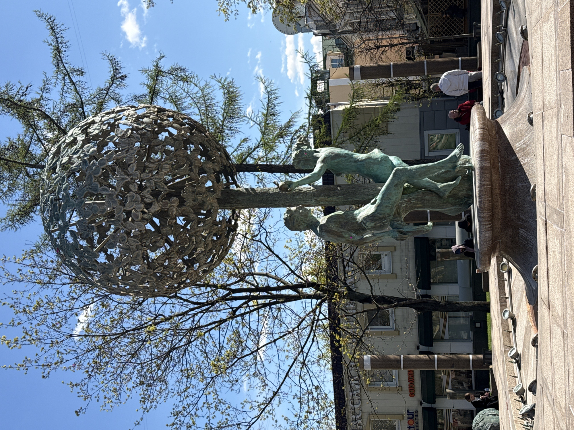 The image captures a bronze statue in an urban setting. The statue features two figures, one reclining and the other reaching towards a sphere-shaped sculpture constructed of leaves. A tree with bare branches partially obscures the statue and the sphere.
In the background, buildings line the street, some with store signs. Two people are visible on the edge of a stone structure, one wearing a red jacket and another in white. The sky is blue with scattered clouds.