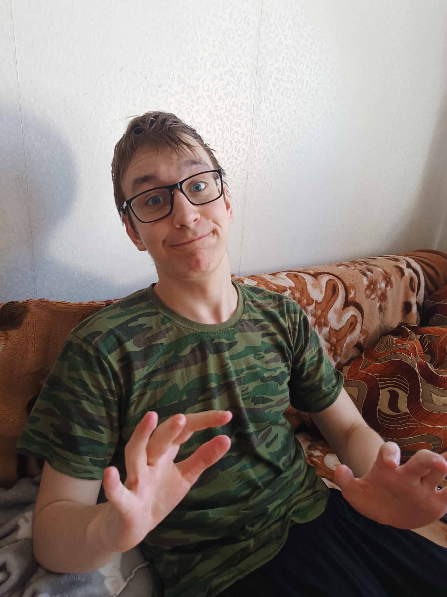 A young man with brown hair and glasses sits on a patterned couch against a white patterned wall. He is wearing a green camouflage t-shirt and black pants. He is holding his hands up, palms facing the viewer, fingers slightly curled. The location is Unknown, Unknown.