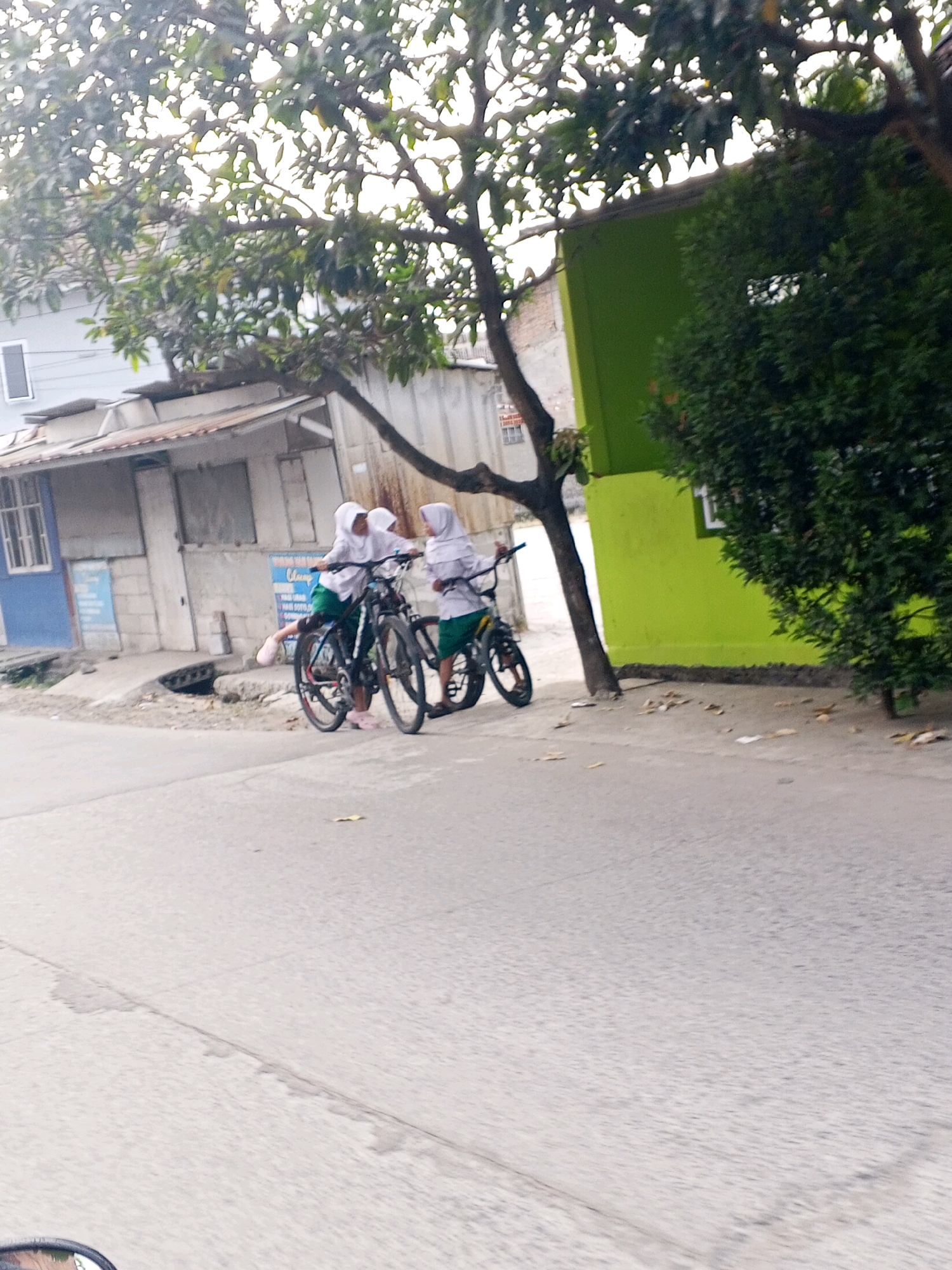 In the city of Ciruas, Indonesia, two young girls in hijabs and school uniforms stand with bicycles on a paved road beside a building with a bright green facade. The girl on the left, wearing a white hijab and green skirt, is seated on a bicycle, her left foot on the ground. The girl on the right, also in a white hijab and green skirt, stands next to her bicycle. Both bicycles are dark-colored and appear to be mountain bikes. Behind them, a street-side building with a corrugated metal roof and blue trim is visible, with a tree branching over the scene. The ground is a rough, paved surface with some debris scattered around. The sky is overcast.