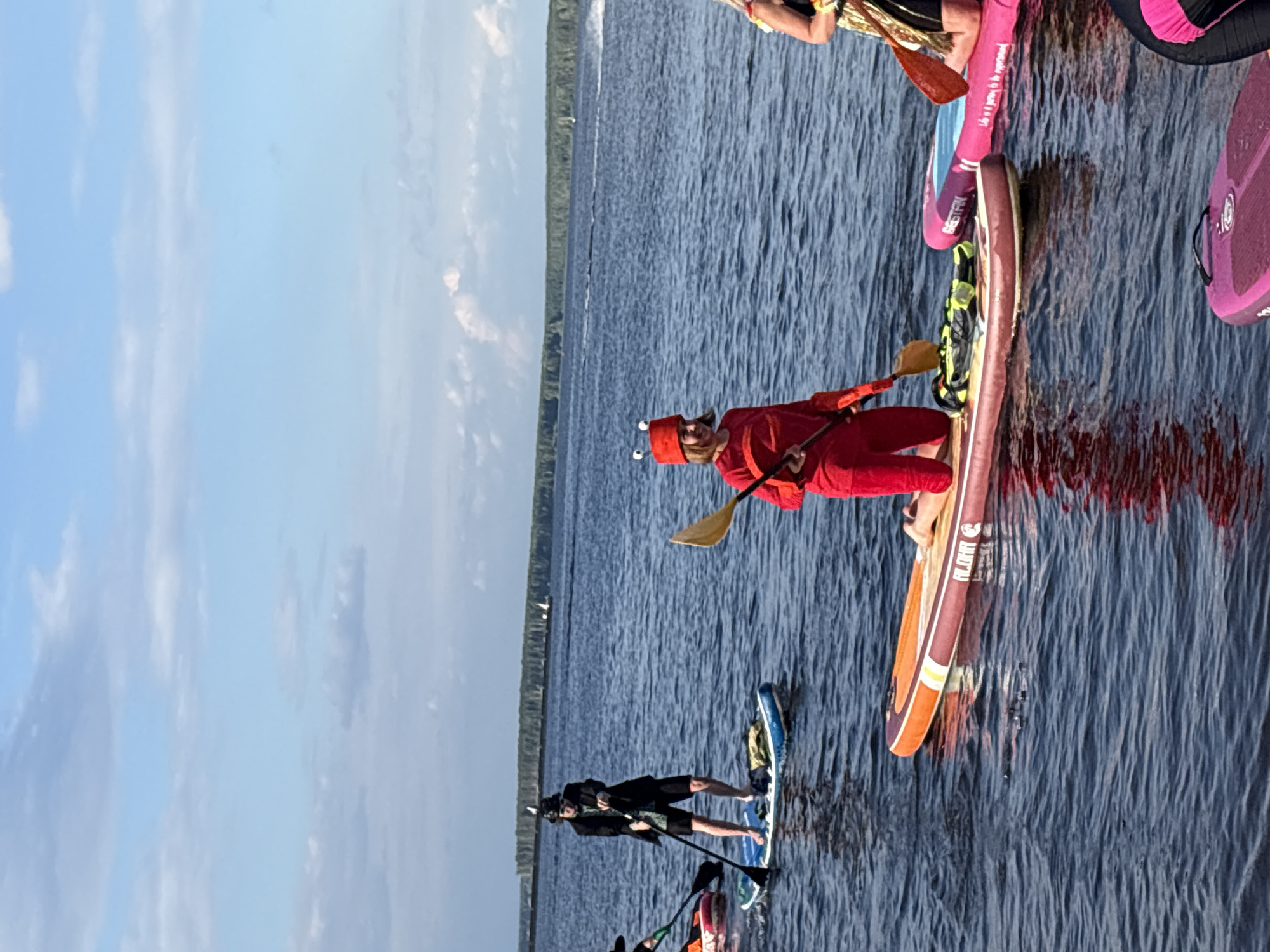 A group of people are paddleboarding on a calm body of water under a bright sky with scattered clouds, suggesting a pleasant daytime. The water appears to be a lake or a wide river, with a tree-lined shore visible in the distance. The overall mood seems to be relaxed and recreational, with individuals enjoying the activity.
One person in the foreground is wearing a red costume with a hat featuring two eye-like attachments, possibly a lobster costume, and is holding a paddle. Another person is further back on the water, dressed in black shorts and a black top, also with a paddle. Several paddleboards are visible, in various colors including pink, purple, blue, and orange. One paddleboard has visible text on its side which appears to be "BESTFIT". Another paddleboard has the text "FLIGHT" on its side. The scene depicts an outdoor activity, likely a casual gathering or event, on a clear day.