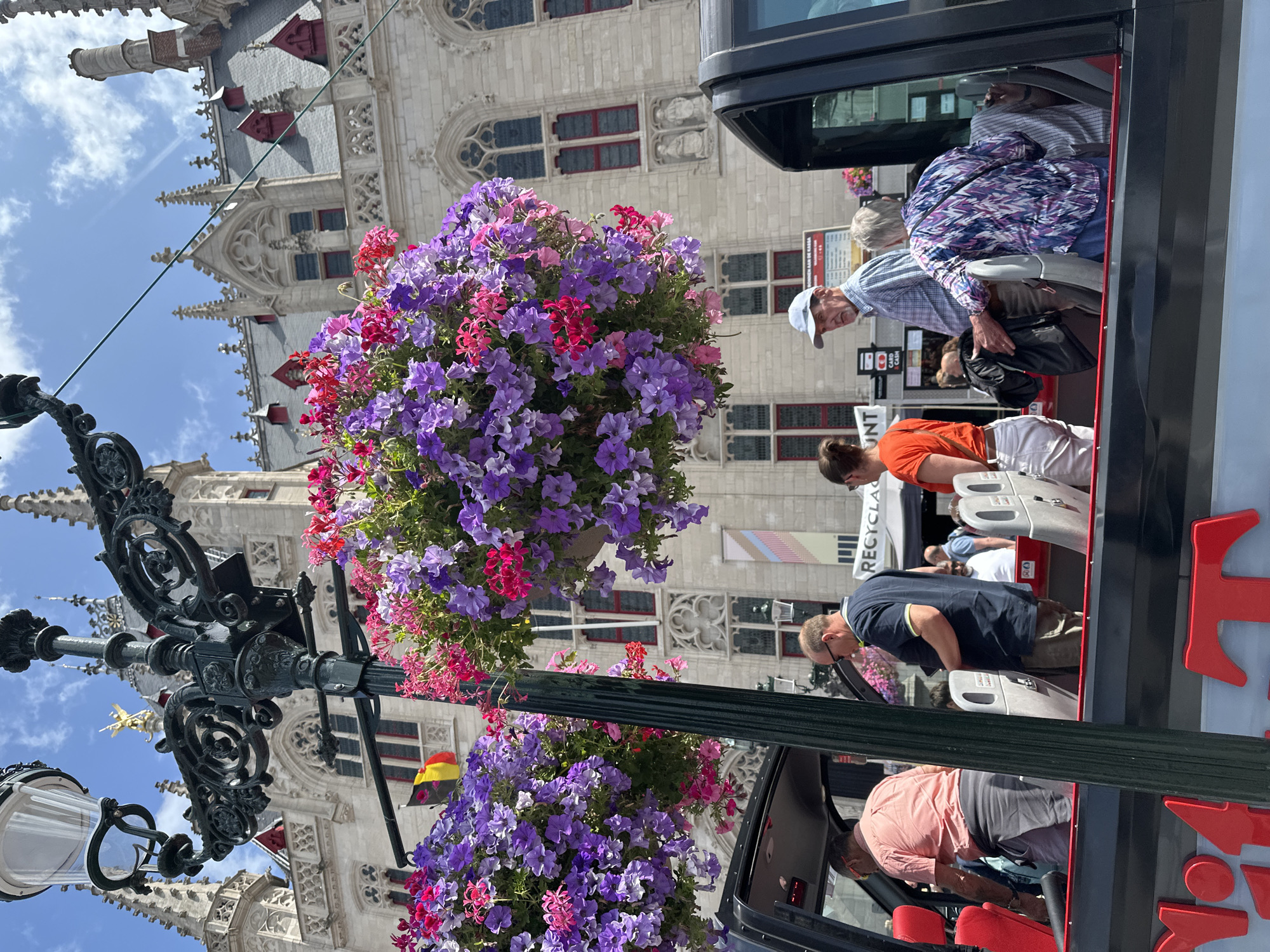 An outdoor, slightly low-angle, medium shot captures a street scene in Bruges, Belgium, on a sunny day with a few scattered clouds. The foreground features the top of a black tour bus, its interior visible with several passengers. A tall, dark green lamppost with ornate wrought-iron detailing stands prominently, holding two large hanging baskets overflowing with vibrant purple and pink flowers.
Behind the lamppost and flowers, a light-colored stone building with Gothic architectural elements, including pointed arches and spires, dominates the background. A Belgian flag, red, yellow, and black, is visible on the building. Several people are seated and standing on the open-top tour bus. On the far left, a man in a coral shirt and grey pants is visible. To his right, a man in a navy blue polo shirt and khaki shorts is looking down. A woman in an orange shirt and white pants stands to the right of him. An older couple is seated in the back of the bus; the woman wears a colorful, patterned top, and the man wears a plaid shirt and a light blue cap. Other passengers are partially visible. Signs are visible behind the passengers on the bus, including one with text that reads "RECYCLAG." The overall scene suggests a tourist attraction in a historic European city.