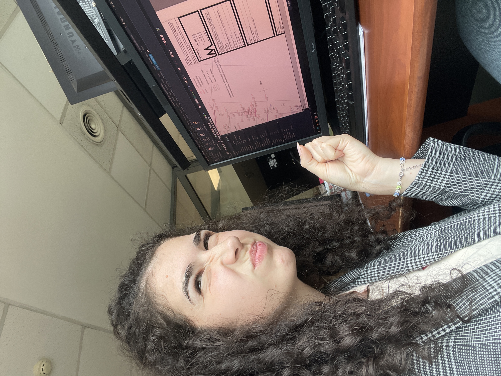An indoor, eye-level shot captures a young woman with dark, curly hair seated at a desk, making a playful "pouty" face with squinted eyes. Her right hand is clenched into a fist and raised, with long, light-colored fingernails visible. She is wearing a grey and white plaid blazer, and a multi-colored beaded bracelet adorns her right wrist.
In the background, a computer monitor displays a technical document or architectural plan, possibly in CAD software, with a predominant pink/reddish color scheme. Cyrillic text is visible on the screen, indicating Bulgarian language. Text snippets include "работна кота" (working elevation), "проектна кота" (design elevation), "теренна кота" (terrain elevation), "конструктиви" (constructive elements), "насип 500-3м³" (fill 500-3 m³), "дебелина на конструкция 0.25m." (thickness of structure 0.25m), "усилена настилка дебелина на конструкция 0.25m." (reinforced pavement thickness of structure 0.25m), and "декоративни плочи дебелина." (decorative tiles thickness.). The monitor brand "PX2611W" is visible on the casing, and a "HYUNDAI" monitor is partially seen behind it. The document also shows "STUDIO MORPHE," a large 'W' or 'M' graphic, and numbers like "7823" and "42.524."
The setting is an office or workspace, indicated by the desk, computer equipment, and the ceiling, which features white acoustic tiles and a circular air vent. A smoke detector or similar fixture is visible on a nearby wall. The visible text in Bulgarian, coupled with the provided context, suggests the location is in Sofia, Bulgaria. The overall mood is lighthearted and candid, likely a posed selfie or casual photo taken during work.