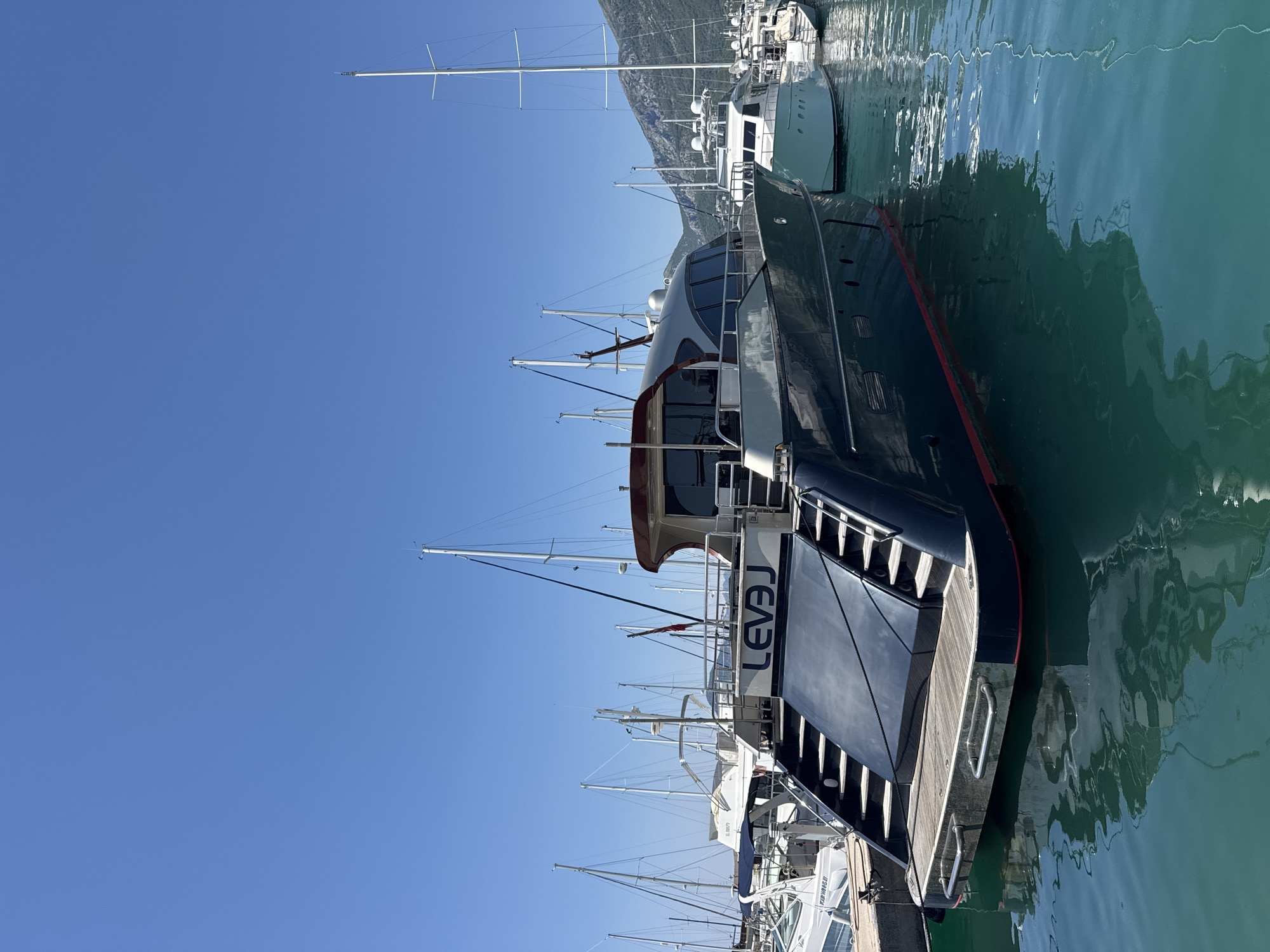 This is a vertically oriented, full-shot image of a dark-hulled yacht named "LEVAJ" docked in a clear blue harbor. The yacht is the most prominent subject, positioned in the foreground and extending towards the right side of the frame. Its sleek, modern design is evident, with large tinted windows and a polished wooden deck at the stern. The name "LEVAJ" is clearly visible in white lettering along the side of the yacht.
Beyond the "LEVAJ," other boats, including sailboats with tall masts, are moored in the harbor. The water is a vibrant turquoise, reflecting the clear blue sky and the surrounding boats. In the background, a mountainous landscape is visible under a bright, cloudless sky, suggesting a sunny day. The scene appears to be a marina or harbor, likely in a coastal town or city.
The image conveys a sense of calm and luxury, characteristic of a serene day at a yachting destination. The clear sky and calm water indicate pleasant weather, possibly midday given the bright sunlight. The location context of Bar, Montenegro, aligns with the presence of a marina and the mountainous backdrop. No people are visible in the frame.