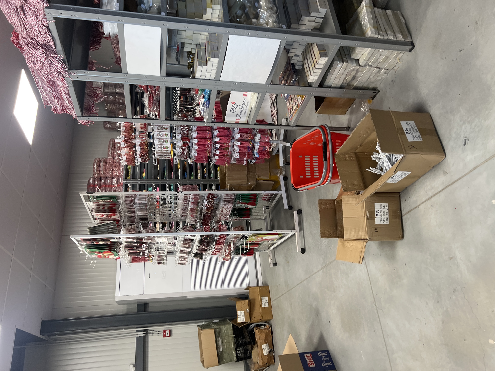 The image depicts the interior of a warehouse or storage facility in Burgas, Bulgaria. The room is filled with tall metal shelving units stocked with various merchandise, primarily holiday ornaments and decorative items. The items are arranged on shelves and hung on racks. Several open cardboard boxes are scattered on the concrete floor, some empty and some containing small white objects, possibly ornament hooks or clips. A bright orange plastic shopping basket sits near the boxes. The space appears to be a storage area for a retail or wholesale business, and the atmosphere is utilitarian, suggesting a functional space for inventory management. A fire alarm system is visible on the wall. The packaging in one of the boxes has the label "Rose Impex Cosmetic Company".