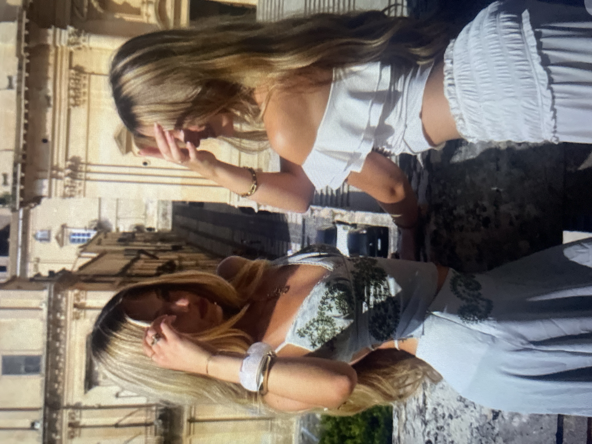 Two young women with blonde hair are posing for a photo outdoors. The woman on the left is wearing a light blue dress with a floral pattern and is touching her forehead. The woman on the right is wearing a white off-the-shoulder top and a white skirt, and she is also touching her forehead. They are standing on a stone balcony overlooking a city with old buildings. The overall lighting suggests it is a sunny day. There are no recognizable landmarks to indicate the location is Kolobrzeg, Poland.