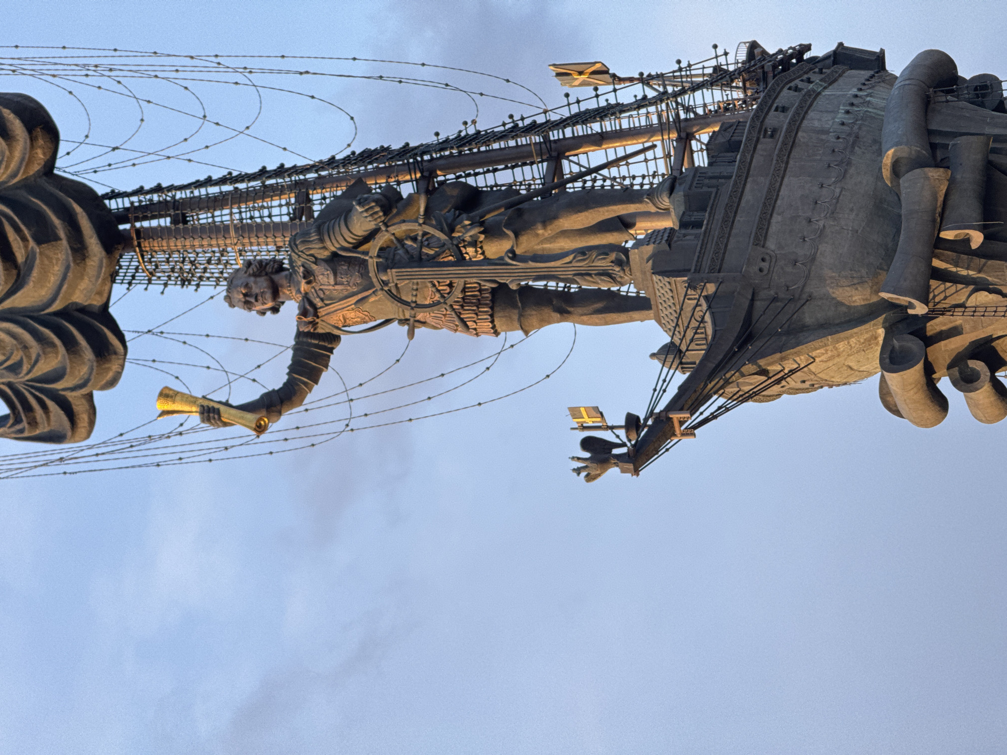 The image shows a close-up view of the Peter the Great Statue in Moscow, Russia, against a light blue sky. The statue depicts Peter the Great holding a golden scroll in one hand and gripping the helm of a ship with the other. The ship is adorned with rigging and sails. The sculpture appears to be made of dark metal or bronze. The image is taken from a low angle, emphasizing the statue's height and scale.