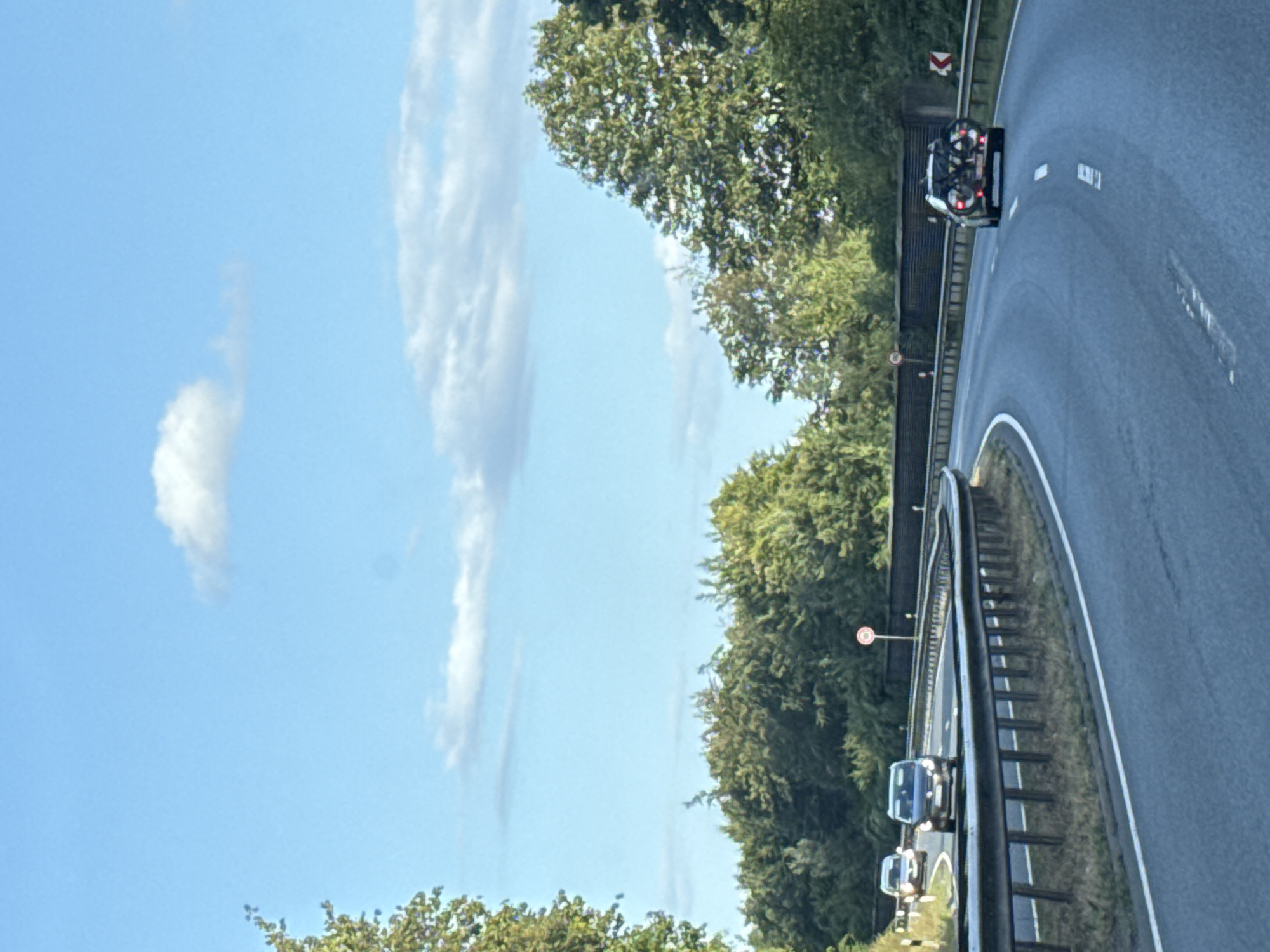 The image shows a road in Herzberg, Germany, with several cars driving on it. The road is a two-lane highway with a guardrail separating it from the oncoming traffic. The cars are all moving in the same direction, and the weather appears to be clear with a few clouds in the sky. There are trees and vegetation on either side of the road, and a speed limit sign of 100 is visible. One car in the foreground appears to have a bicycle rack on the back.