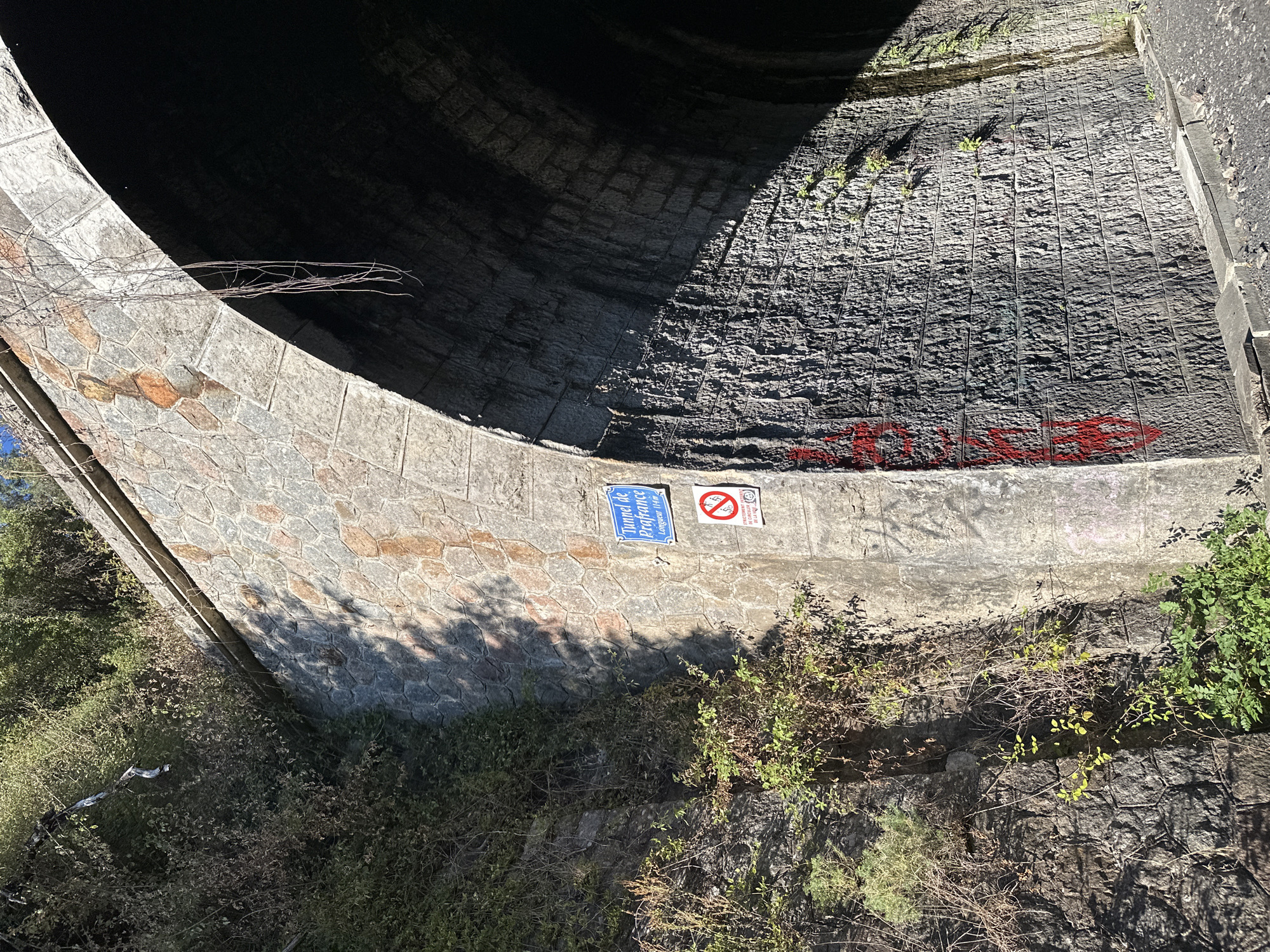 The image shows the exterior of a stone tunnel entrance, identified by a blue sign as "Tunnel de Prafrance," with a length of 114 meters. The tunnel is surrounded by overgrown vegetation and trees. A white sign with a red circle and a diagonal line through a bicycle symbol indicates that cycling is prohibited. Red graffiti is visible on the stonework near the tunnel entrance. The scene appears to be in Générargues, France.