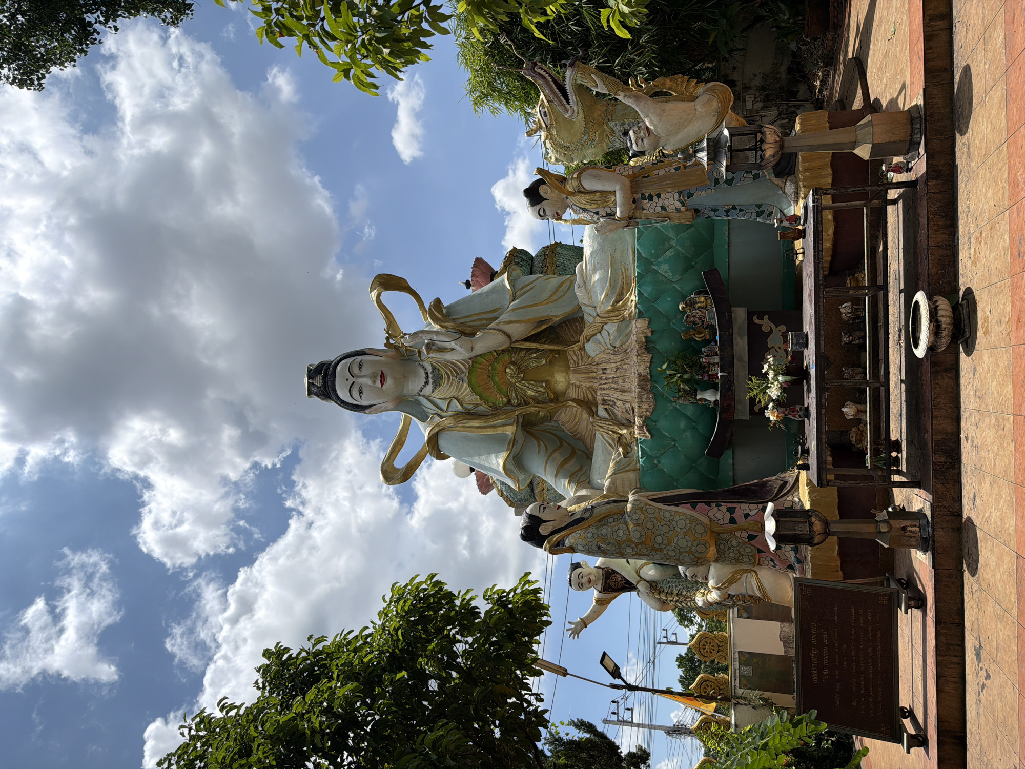 The image displays a large, ornate statue of a seated deity, likely Guan Yin, against a bright blue sky with scattered white clouds. The deity statue is adorned with flowing robes and elaborate golden headdress, and its hands are raised in a gesture of blessing. Flanking the main deity are smaller statues of figures in traditional attire. To the right of the main statue, a mythical dragon-like creature is depicted. The statues are situated on a raised platform covered with patterned tiles. In front of the platform, a metal table holds offerings and decorative items, and a stone urn is placed on the ground. A sign with Thai text is visible to the left of the platform. The surroundings are lush with green foliage, suggesting an outdoor location. This scene is located at the Wat Pha Lat temple in Chang Phueak, Thailand.