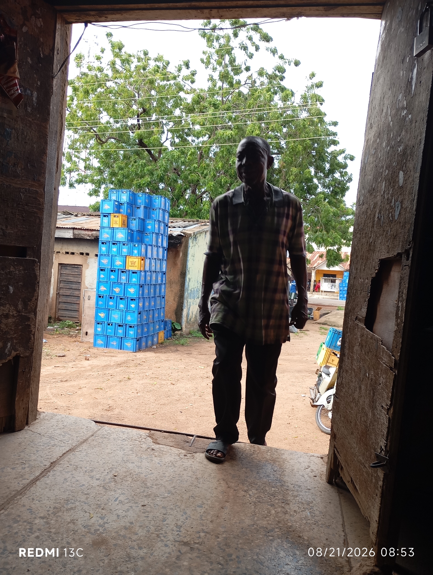 A man stands in a doorway, facing the camera. He is wearing a checkered shirt and dark pants, with sandals on his feet. Behind him, a large stack of blue crates is visible, along with some buildings and trees. The ground is dirt, and there is a motorcycle parked to the right of the frame. The image appears to be taken in an outdoor market or storage area.