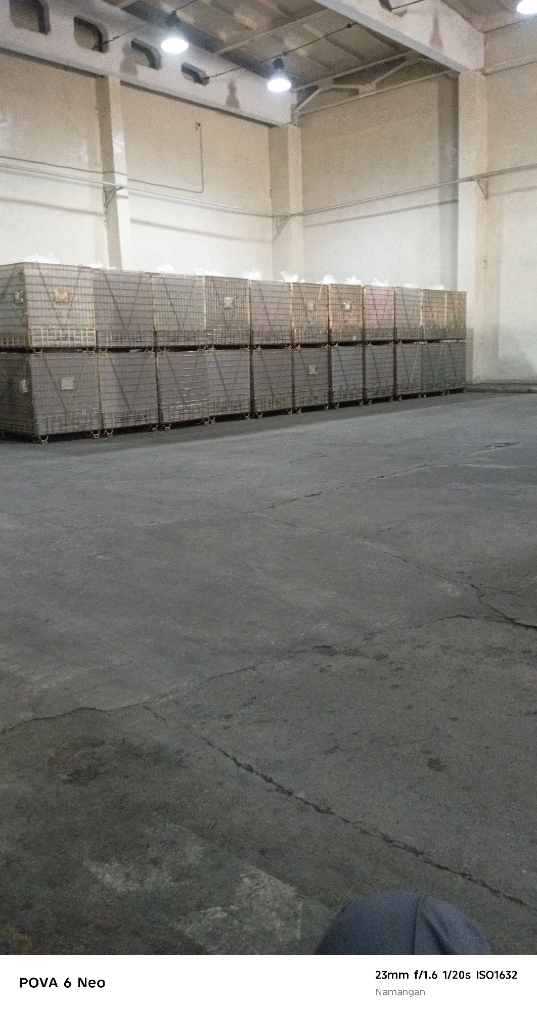The image shows a dimly lit warehouse interior with large stacks of metallic cages filled with what appear to be wrapped goods. The cages are arranged in two rows, one stacked on top of the other, extending horizontally across the frame. The warehouse has a high ceiling with exposed beams and industrial lighting fixtures. The floor is a concrete surface, showing signs of wear and cracking. In the foreground, a small portion of a person's leg, clad in dark fabric, is visible, suggesting a person is present, possibly the photographer. The text overlay in the bottom right corner indicates the location as Namangan, Uzbekistan, and provides camera settings: 23mm f/1.6 1/20s ISO1632.