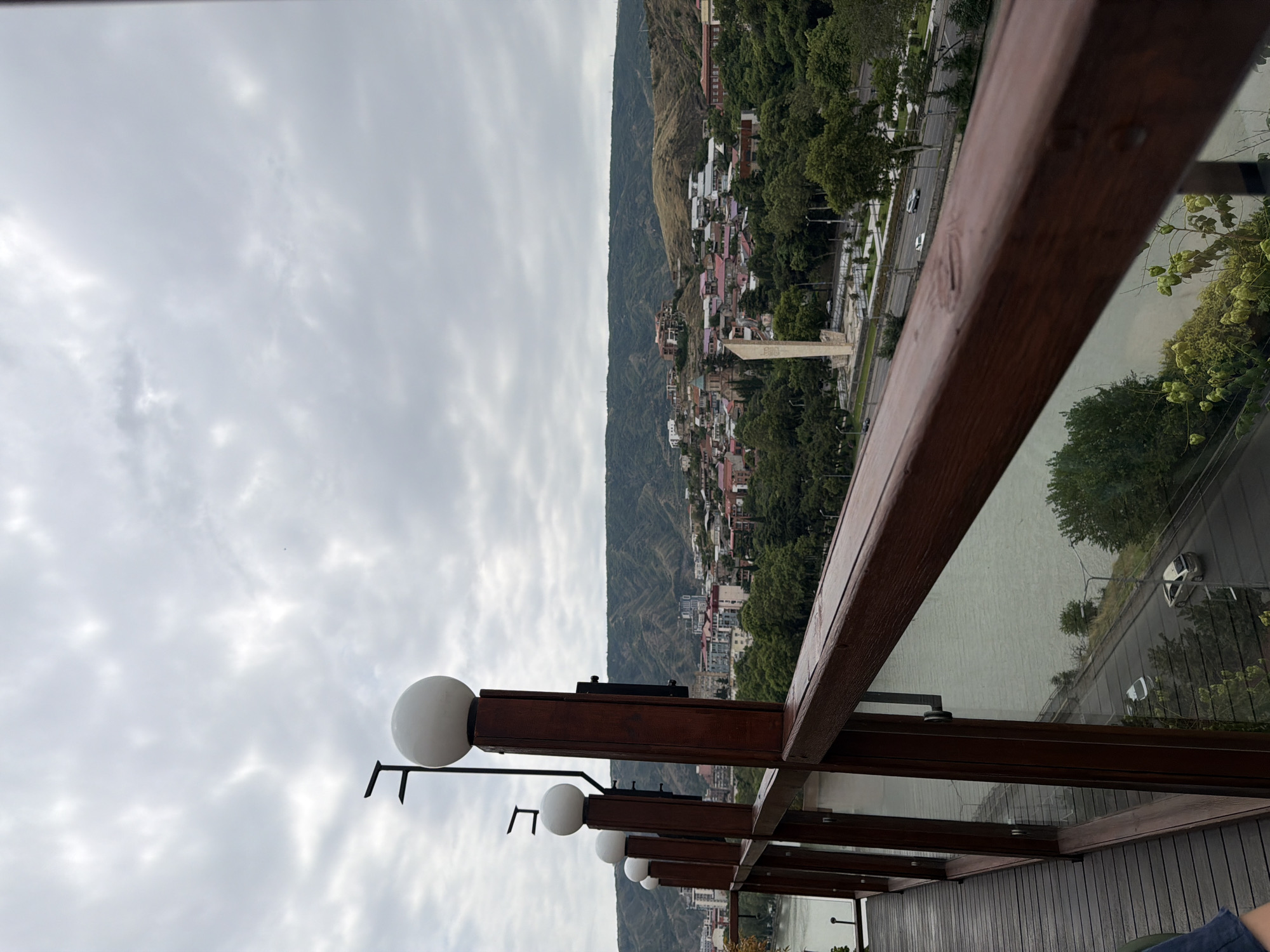 This is a vertical, eye-level shot of a city view of Tbilisi, Georgia, seen from a wooden balcony with a railing. In the foreground, a dark brown wooden railing runs diagonally from the bottom right to the middle left of the frame. On top of the railing posts, there are four spherical white lights, three of which are clearly visible. The balcony floor is made of dark wood planks.
Beyond the railing, a wide, murky green river flows from the bottom left towards the middle of the frame. A road runs along the riverbank, with several cars visible. Across the river, the city of Tbilisi unfolds. Numerous buildings with red-tiled roofs and white or light-colored walls are nestled among lush green trees. In the background, rolling hills covered in vegetation rise up, forming a backdrop for the city. A tall, light-colored monument with a pointed top stands prominently in the middle ground, near the river. The sky is overcast with a thick layer of gray clouds. The overall lighting suggests an overcast day.